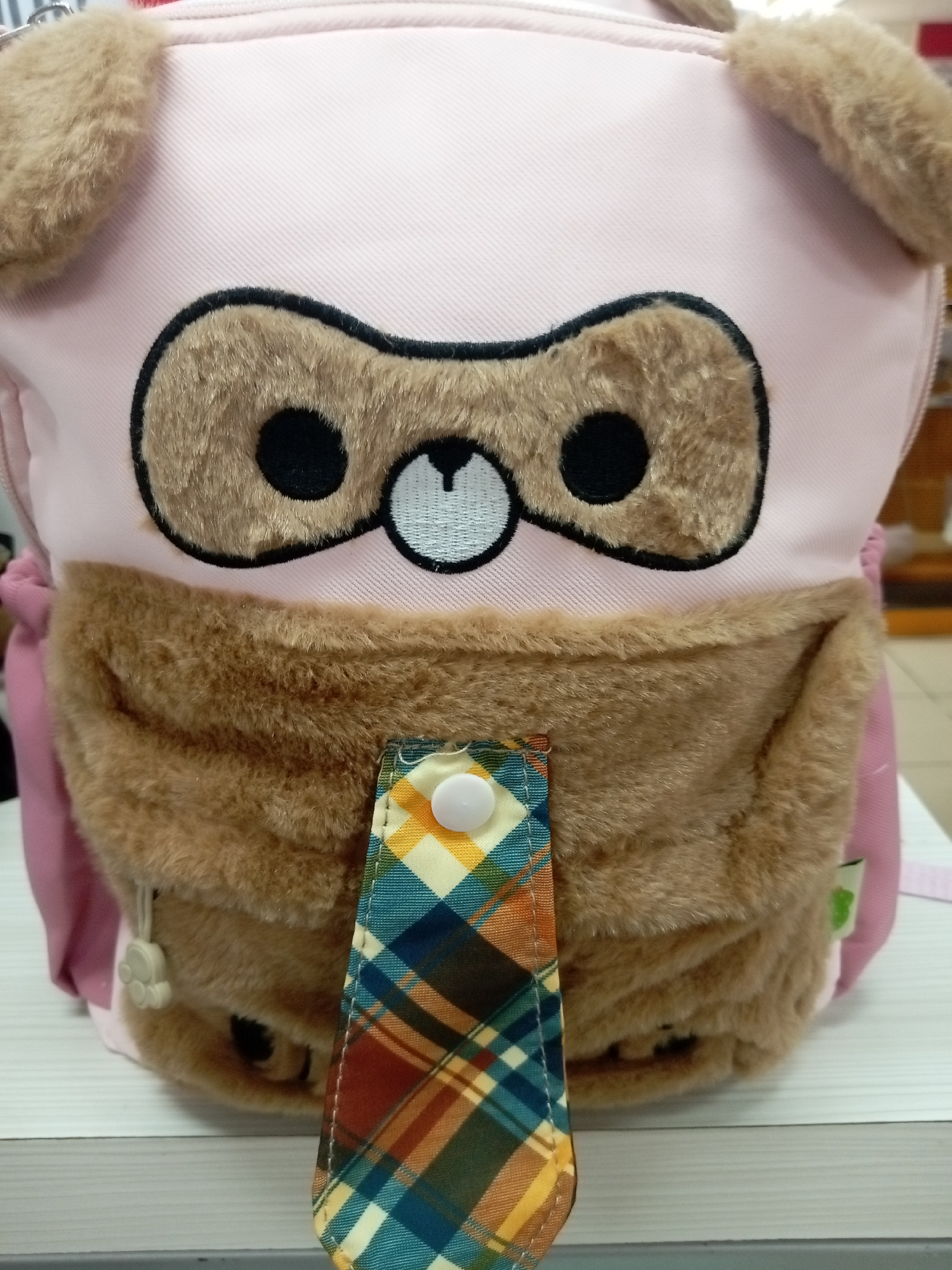 A close-up, frontal view of a pink child's backpack designed to resemble a bear's face. The backpack features plush brown material for the ears, eyes, nose, and mouth area. The eyes are black circles within fuzzy brown outlines, and the nose is a small white embroidered detail. A textured brown pouch is located below the face. Attached to the pouch is a strap with a plaid pattern in shades of blue, yellow, orange, and green, secured by a white button snap. The backpack appears to be resting on a white surface, possibly a table or shelf. The background is blurred and shows hints of a retail environment, suggesting the image was taken in a store. The location of Batam, Indonesia, is not discernible from the image itself.