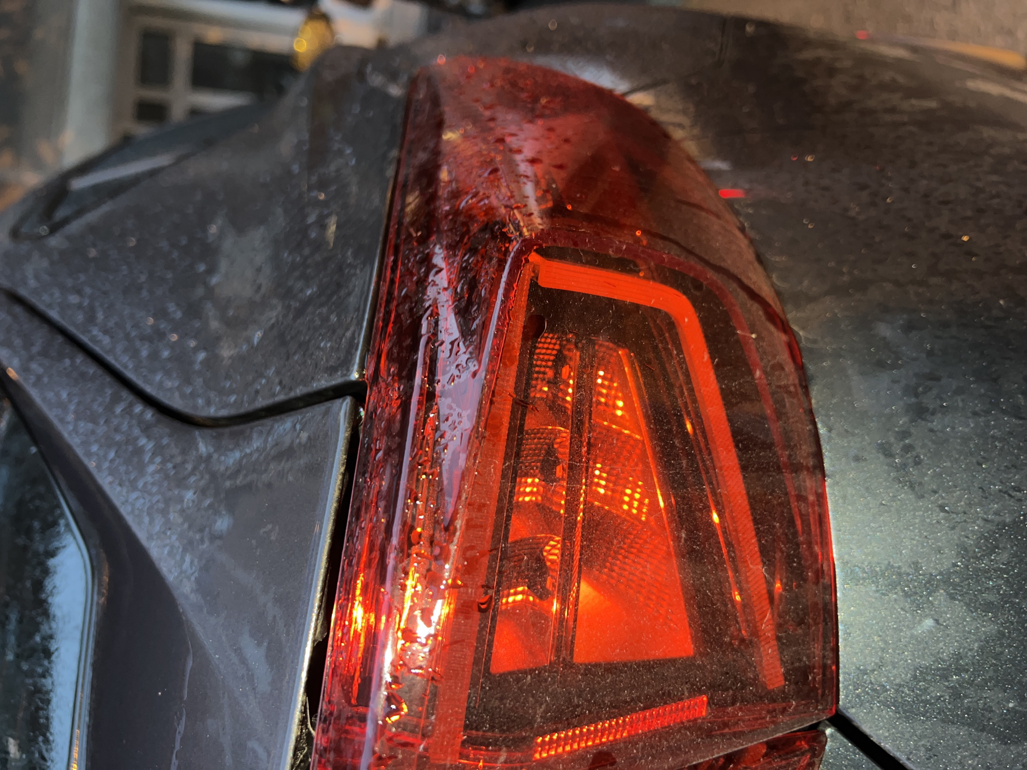The image shows the rear taillight of a gray car. The taillight is red and features a geometric design. Water droplets are visible on the surface of the taillight. The car appears to be dirty, as evidenced by the speckled surface on the car body. The location is not discernible from the image.