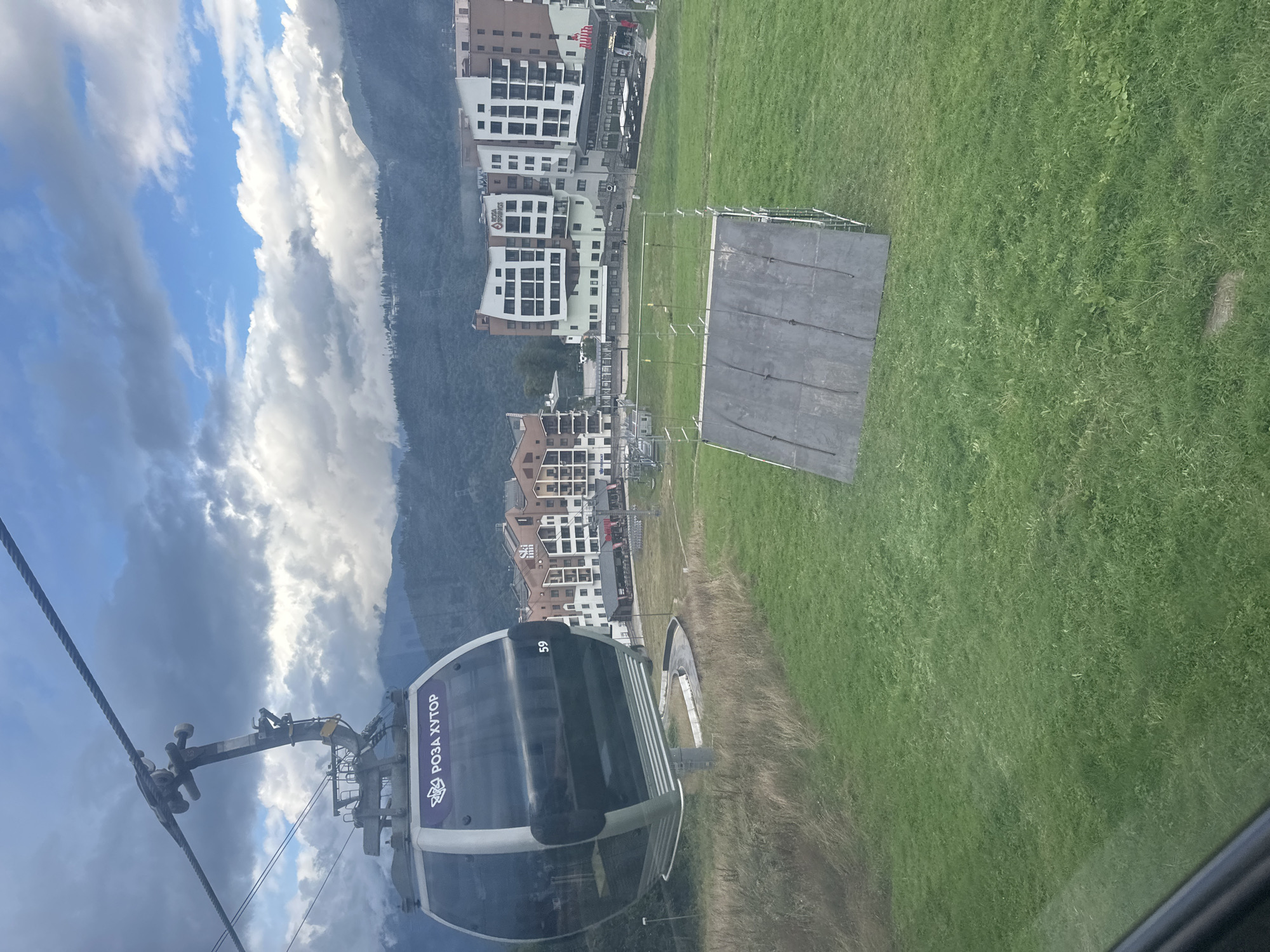 The image depicts a view from a cable car traveling over a grassy hillside in Estosadok, Russia. In the foreground, the cable car itself is visible, labeled with the Cyrillic text "РОЗА ХУТОР" (Rosa Khutor) and the number "59". A ramp-like structure made of metal and dark material sits on the green grass, possibly for a sporting event or activity. Beyond the ramp, several multi-story buildings, likely hotels or resorts, are arranged in a village-like cluster. The buildings feature a mix of white, brown, and beige facades with numerous windows. In the background, misty mountains rise, partially obscured by clouds. The sky is a mix of blue patches and heavy gray clouds, suggesting variable weather conditions. The overall scene conveys a mountainous resort environment.