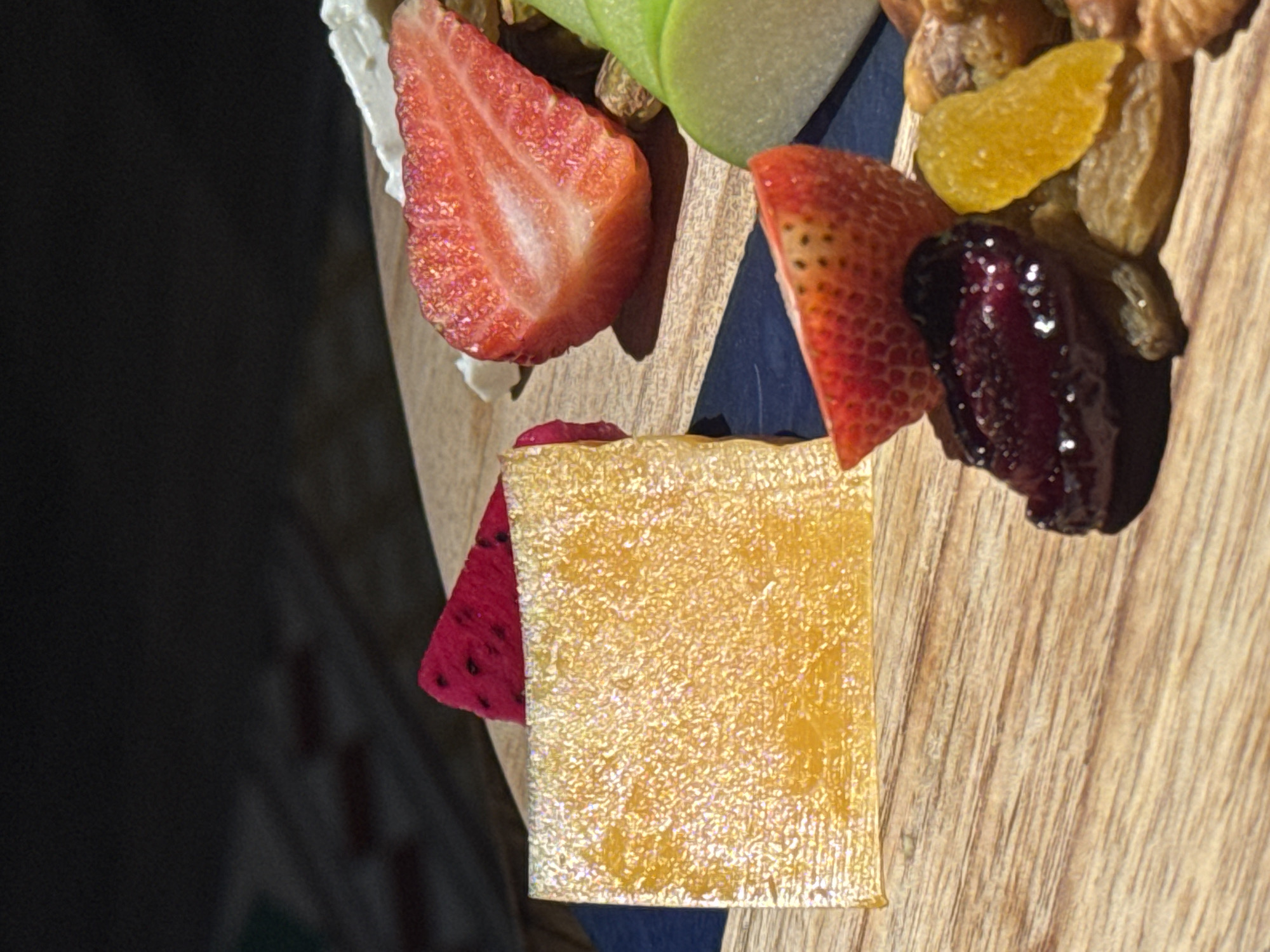 A close-up shot showcases a colorful array of fruits and sweets on a wooden board. In the foreground, a square piece of translucent, golden jelly rests next to a slice of strawberry and a pile of dried fruits, including raisins and what appear to be dried cranberries and apricots. Behind these, a vibrant pink slice of dragon fruit is visible, partially obscured by the jelly. Further back, a half strawberry sits atop a dollop of white cream, and slices of green apple are arranged to the right. The background is a dark, out-of-focus area, suggesting an indoor setting with dramatic lighting that casts shadows. The overall impression is one of a carefully arranged dessert or snack platter. There is no information within the image to determine its location in Hà Nội, Vietnam.