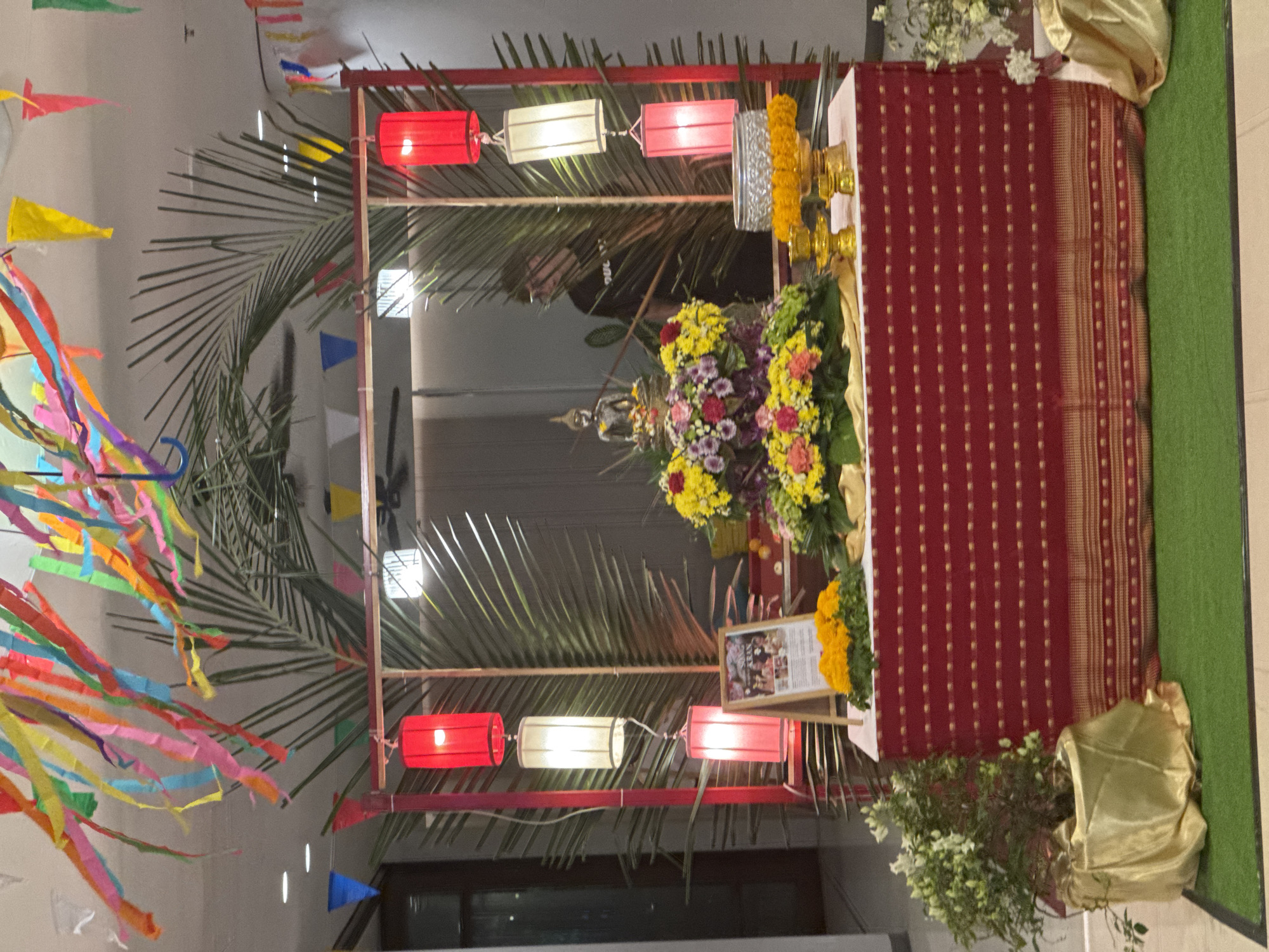 The image captures a small, makeshift Buddhist shrine indoors in Patong, Thailand. The shrine is constructed with a simple wooden frame adorned with red and white paper lanterns and palm fronds. Strings of colorful pennants and ribbons are draped on the left side of the frame.
At the center of the shrine, a seated Buddha statue is partially visible amidst a colorful arrangement of fresh flowers, including yellow chrysanthemums and purple and pink blooms. Behind the flowers, an ornate silver and gold offering tray is placed. To the right of the shrine, a table covered with a red cloth with gold embellishments serves as a backdrop.
A framed photograph is positioned on the table in front of the cloth. Below the table, a piece of golden fabric is draped. At the base of the shrine, a pot of white flowers is placed. The scene is illuminated by overhead lighting.