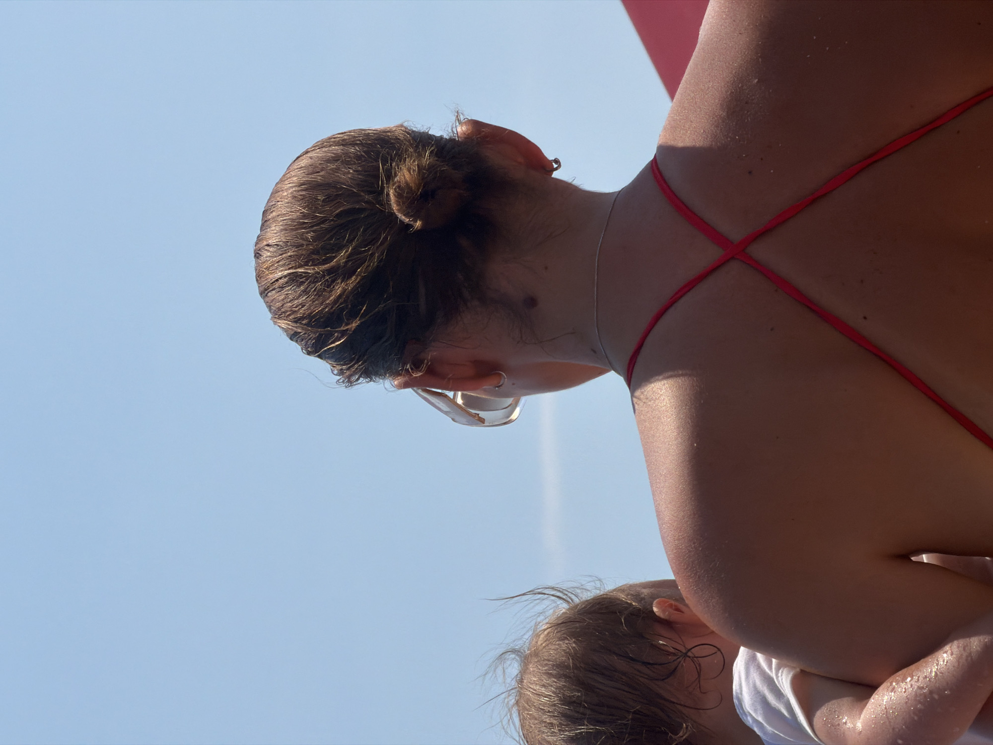 An overhead, outdoor, medium shot captures the back of a woman as she holds a baby. The woman's dark hair is pulled into a bun at the nape of her neck, and she wears large, light-brown sunglasses. Her back is bare, showing a dark red, crisscrossed strap bikini top. The baby is partially visible, with its back to the camera and its head turned to the side. The background is a clear blue sky. The scene appears to be a sunny day in Budva, Montenegro.