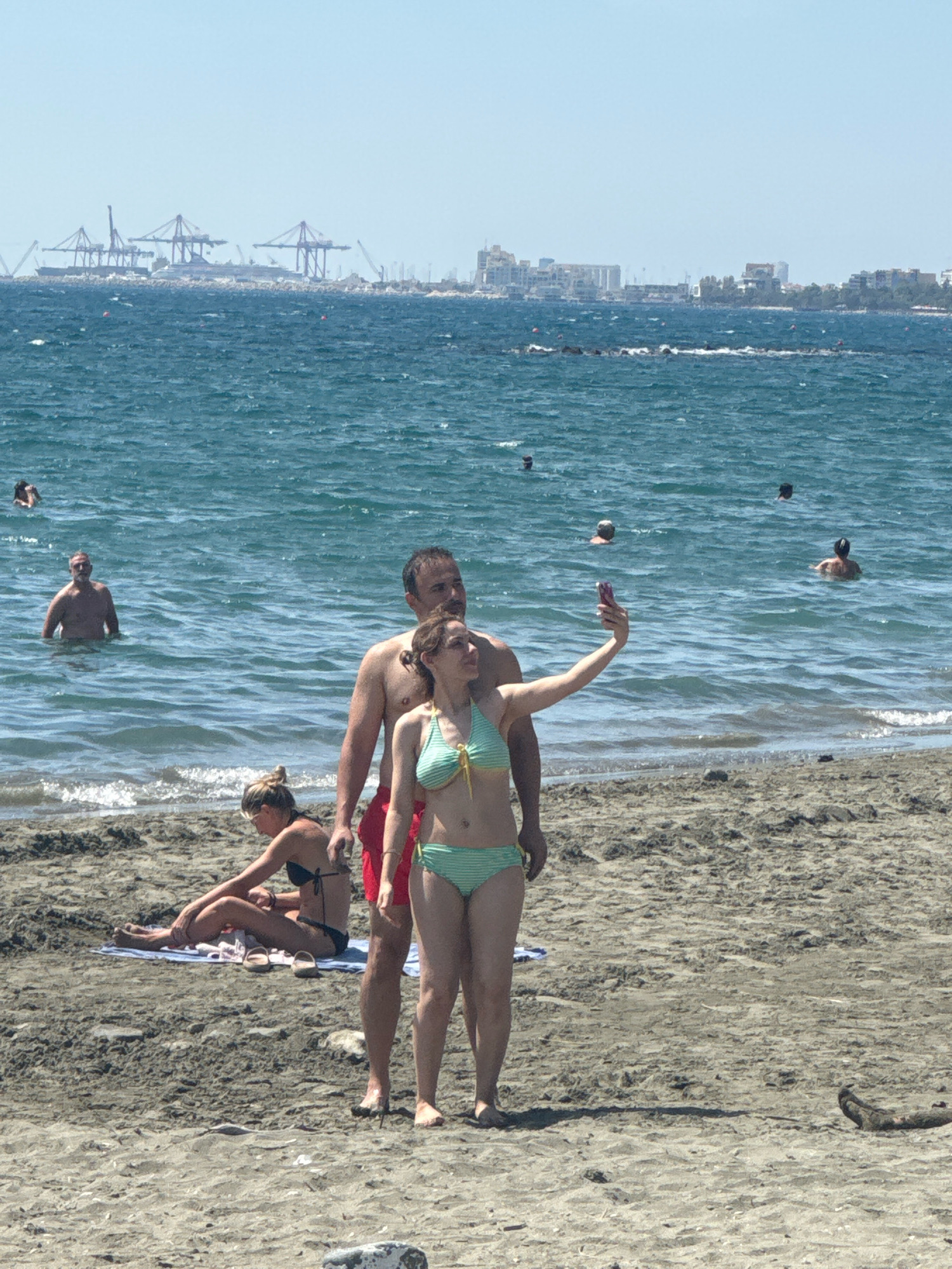 The image depicts a sunny day at a beach, likely in the Germasogeia Municipality, Cyprus, given the visible cityscape and port infrastructure in the background. In the foreground, a couple is taking a selfie by the water. The woman, wearing a green and white striped bikini, holds a pink smartphone up to capture the photo. The man, in red swim trunks, stands next to her. Behind them, a woman in a black bikini sits on a towel on the sand, facing away from the camera. Further out in the ocean, several individuals are swimming and wading. The sea is a vibrant blue with moderate waves. The distant coastline features buildings and numerous cranes, indicating a port or industrial area. The beach itself is composed of dark sand with scattered small stones and debris. The sky is clear and bright.