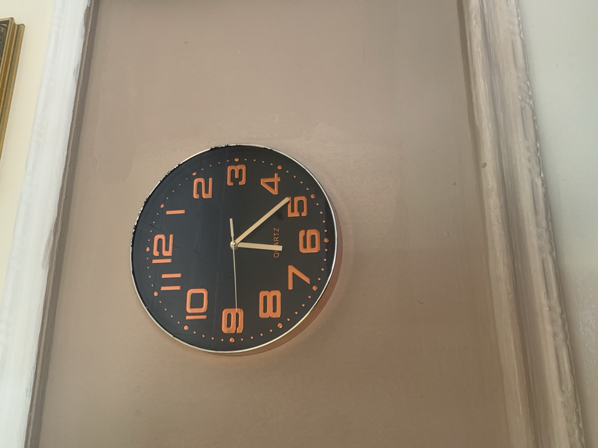The image shows a close-up of a round, black wall clock with orange numerals, mounted on a beige-colored wall. The clock has two hands; the hour hand points to approximately 10, and the minute hand points to approximately 4. The word "QUARTZ" is visible below the center of the clock face. A white, decorative molding runs along the bottom of the wall, and the top edge of the wall is also visible, showing a white painted surface. The lighting suggests an indoor environment. The location of Gura Top, Nigeria is not discernible from the image.
