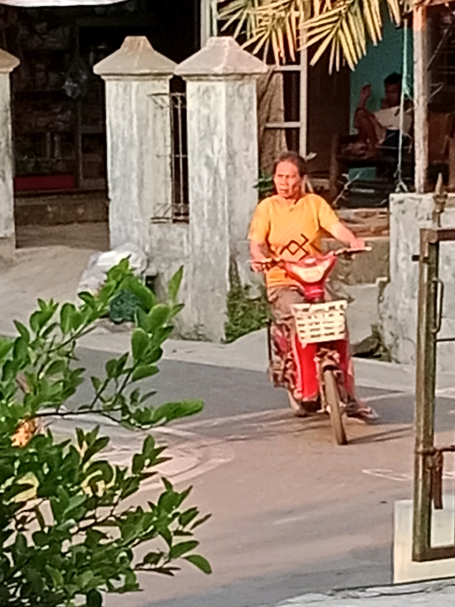 A person wearing an orange t-shirt and a patterned skirt rides a red scooter with a white basket on the front. The scooter is on a paved road. In the background, there are two stone pillars and a building with a window. There are also green leaves in the foreground, partially obscuring the view of the scooter. The image appears to be taken in Waru, Indonesia.