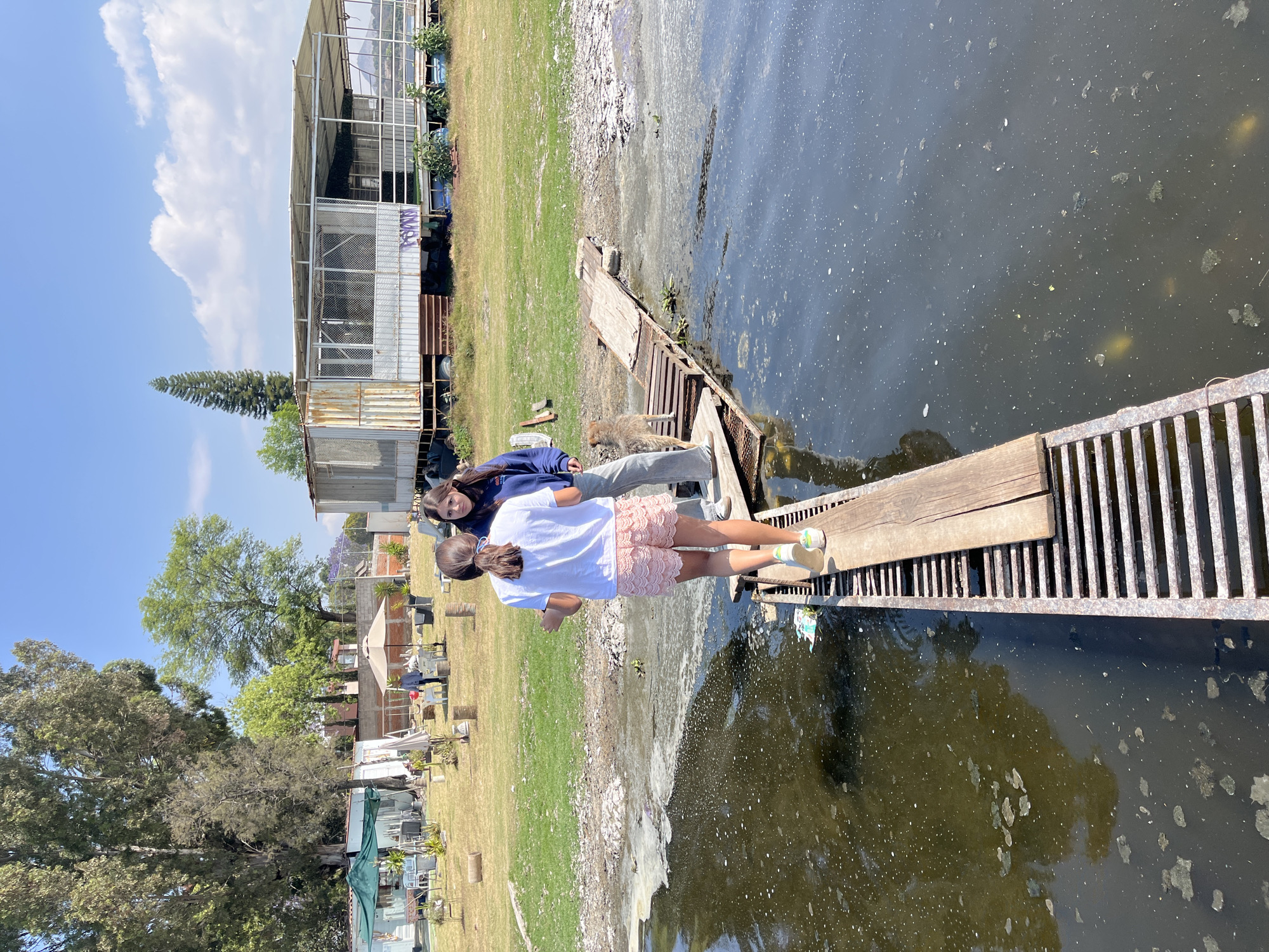 Two young females stand on a metal grate with wooden planks extending over a murky body of water in El Oasis Valsequillo, Mexico. The younger girl, facing away, wears a white shirt and pink lace shorts. The other girl, standing beside her, is older and wears a blue sweatshirt and grey pants, and looks toward the camera. The water is dark and speckled with light-colored debris. The shoreline is visible behind them, with a mix of grass, debris, and a concrete structure. Beyond the shoreline, there are trees and buildings. A blue sky with scattered clouds is above.