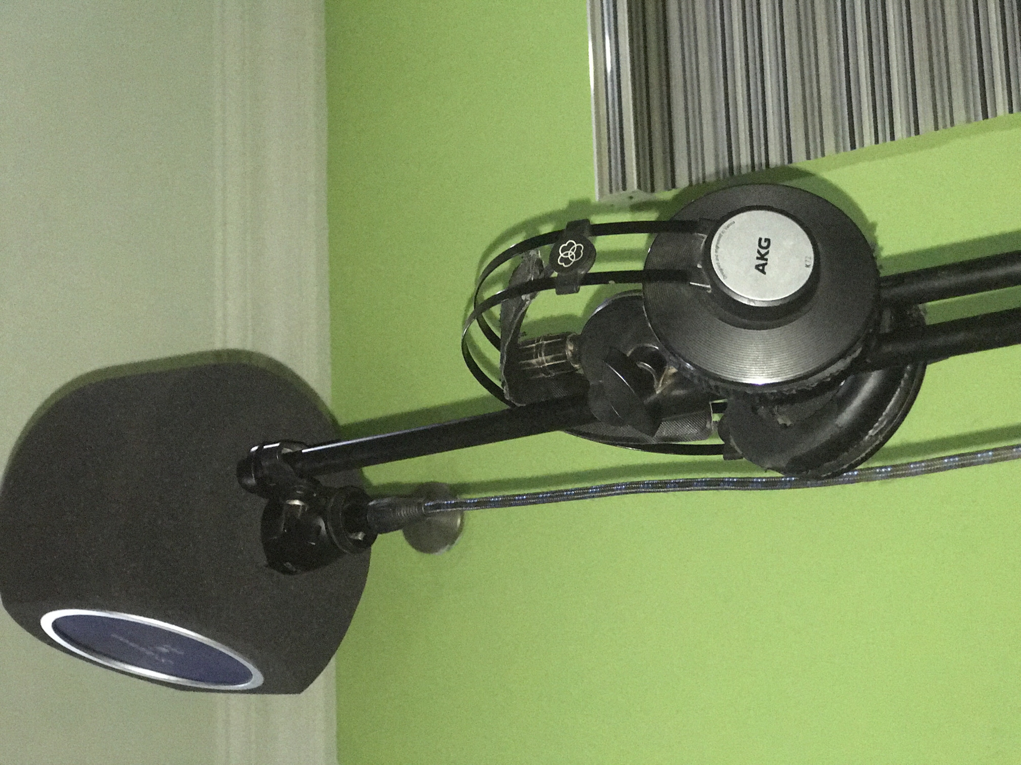 The image displays an indoor audio recording setup, devoid of people. The central focus is a black studio microphone with a large foam pop filter, positioned on the left side of the frame. The microphone's front reveals a silver ring enclosing a dark blue or black interior. This microphone is mounted on a black boom arm that extends horizontally across the bright lime green wall.
Hanging from the boom arm on the right are a pair of black over-ear headphones. A prominent feature is the outer face of one earcup, which has a circular silver plate bearing the brand name "AKG" printed vertically in black capital letters. Beneath this, the text "designed and engineered by Harman" is faintly visible. A small black clip, adorned with an abstract white clover-like logo, is attached to the headphone's headband. A braided cable, primarily blue and black, runs along the boom arm and descends towards the bottom right.
The background consists of a vibrant lime green wall. On the far right, above the headphones, a section of vertical blinds with alternating grey and white stripes is visible, suggesting a window. A light-colored wall or doorframe is partially visible on the far left edge of the image.
The arrangement of the equipment indicates a space prepared for audio recording, such as a home studio or podcasting station. No specific activity is actively occurring, and the time of day is not discernible from the indoor setting. While the location context of Abuja, Nigeria, is provided, there are no visual cues within the image itself to confirm this geographical detail.
