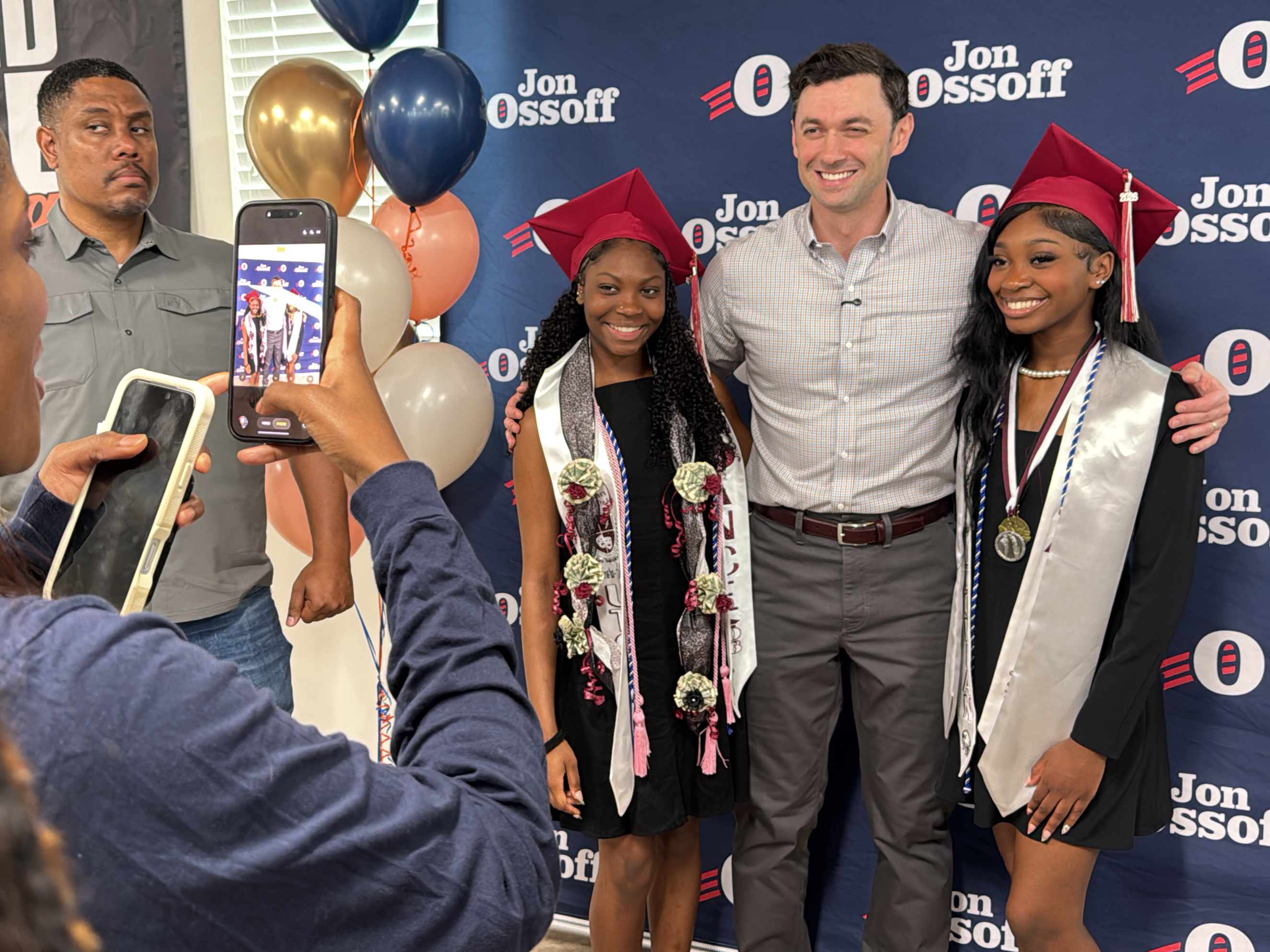 The image captures Jon Ossoff, a U.S. Senator from Georgia, posing with two graduating high school students. They are standing against a backdrop with Ossoff's name and campaign logo. Ossoff, a white man, is wearing a light-colored button-down shirt and grey pants, with his arms around the graduates. The graduates, both young Black women, are wearing graduation gowns and caps. The graduate on the left has a white graduation sash decorated with flowers and money, and the graduate on the right has a similar white graduation sash with a medal. Two adults are partially visible on the left side of the frame, taking photos of the scene with smartphones. Balloons in various colors are visible behind the group. The photo was taken in Decatur, United States.