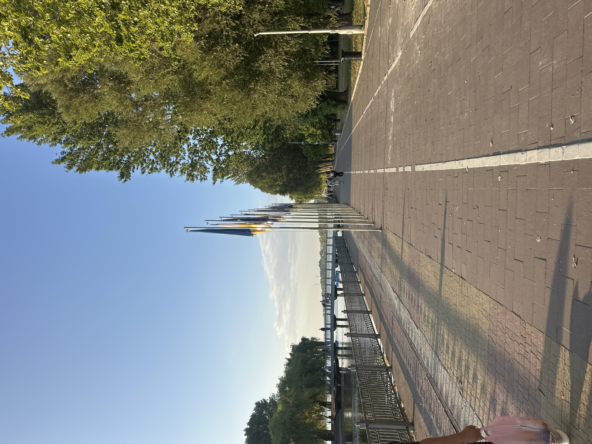 The image depicts a waterfront promenade in Ternopil, Ukraine. A paved walkway, made of brick-like stones, is visible, with a white line dividing it into two lanes, suggesting a path for pedestrians and cyclists. To the left of the walkway is a metal fence separating it from a body of water, likely Ternopil Pond. Beyond the fence, a pedestrian bridge spans the water, with several people walking on it. Trees line the right side of the promenade, casting shadows on the walkway. In the foreground, a person's legs and a pink plastic bag are visible. Several flagpoles with flags are also present along the promenade. The sky is clear and blue, with some clouds.
