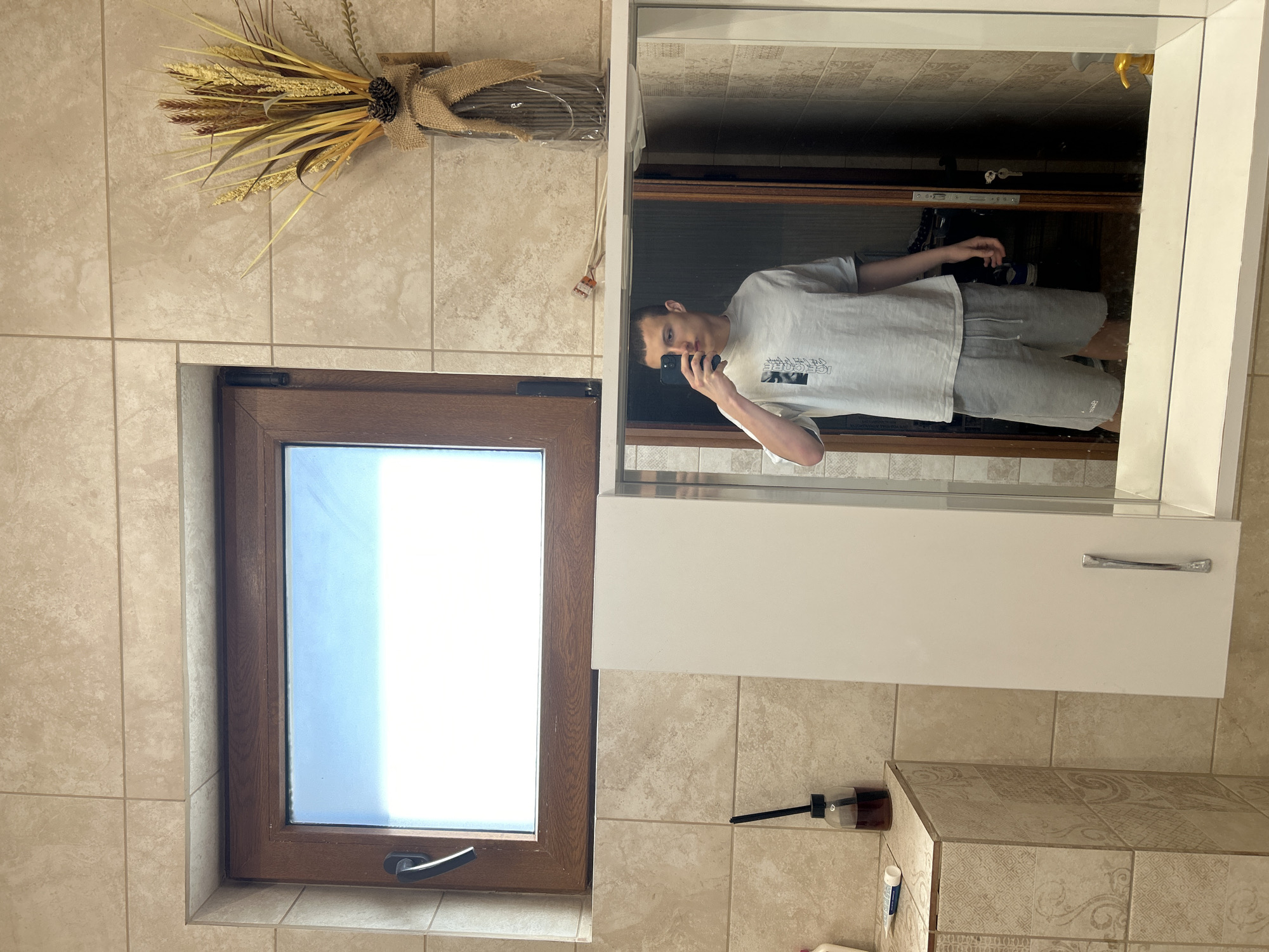A man is visible in a mirror taking a selfie in a bathroom. The bathroom has beige tile walls, a brown-framed window showing a blue sky, and a white cabinet with a mirror. There is a decorative arrangement of dried plants and a pine cone on the wall above the window. A bottle of liquid and a toothbrush are on the countertop. The location is likely in Burgas, Bulgaria.