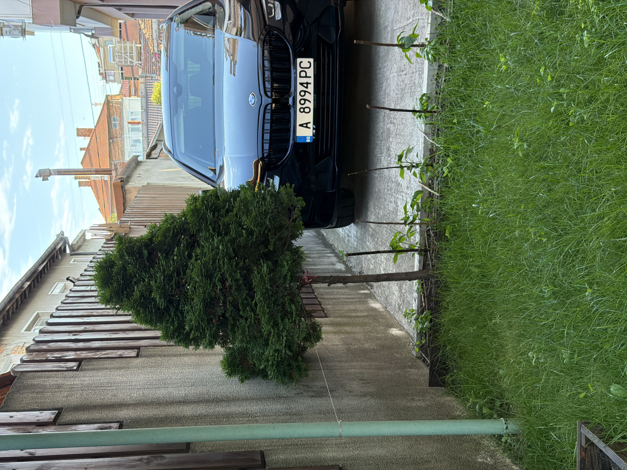 The image depicts a narrow courtyard in a residential area of Burgas, Bulgaria. A black BMW SUV is parked on a concrete surface. To the left of the vehicle, a small, manicured evergreen tree stands in front of a textured concrete wall. The wall is partially covered by vertical wooden planks. Beyond the wall, residential buildings with red tile roofs are visible. In the foreground, a patch of green grass is growing, with several thin wooden stakes supporting small plants. The sky is partly cloudy. The license plate on the BMW reads "A 8994 PC BG".