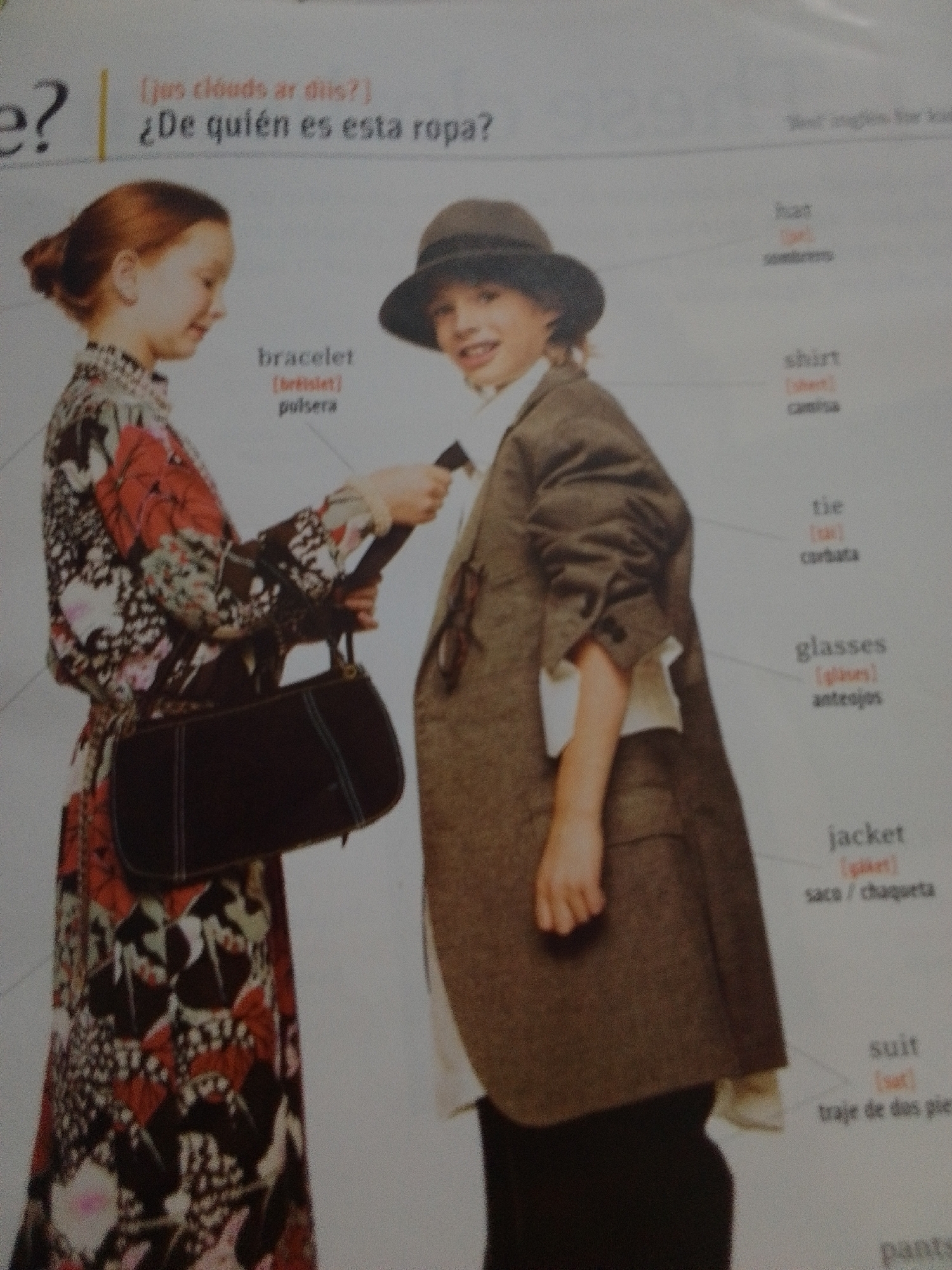 This media file features a close-up shot of two children, a girl and a boy, in what appears to be an educational setting, possibly a classroom or a page from a children's book, given the accompanying text. The girl on the left, with red hair tied back, wears a long-sleeved dress with a vibrant floral pattern in red, pink, black, and white. She is holding a dark, elongated handbag with a strap. The boy on the right, wearing a hat, a jacket, and possibly a shirt, is positioned next to her. The image is accompanied by text in English and Spanish, which seems to be a vocabulary lesson related to clothing. The visible text includes phrases like "¿De quién es esta ropa?" (Whose clothes are these?), "bracelet," "pulsera," "shirt," "tie," "cortata," "glasses," "anteojos," "jacket," "saco/chaqueta," "suit," and "traje de dos pie." The overall tone is educational and informative. The lighting suggests an indoor environment. There are no strong cues about the time of day or weather, nor specific location details beyond the general context of an educational resource. The emotions displayed are neutral, as the children are posing for the image.