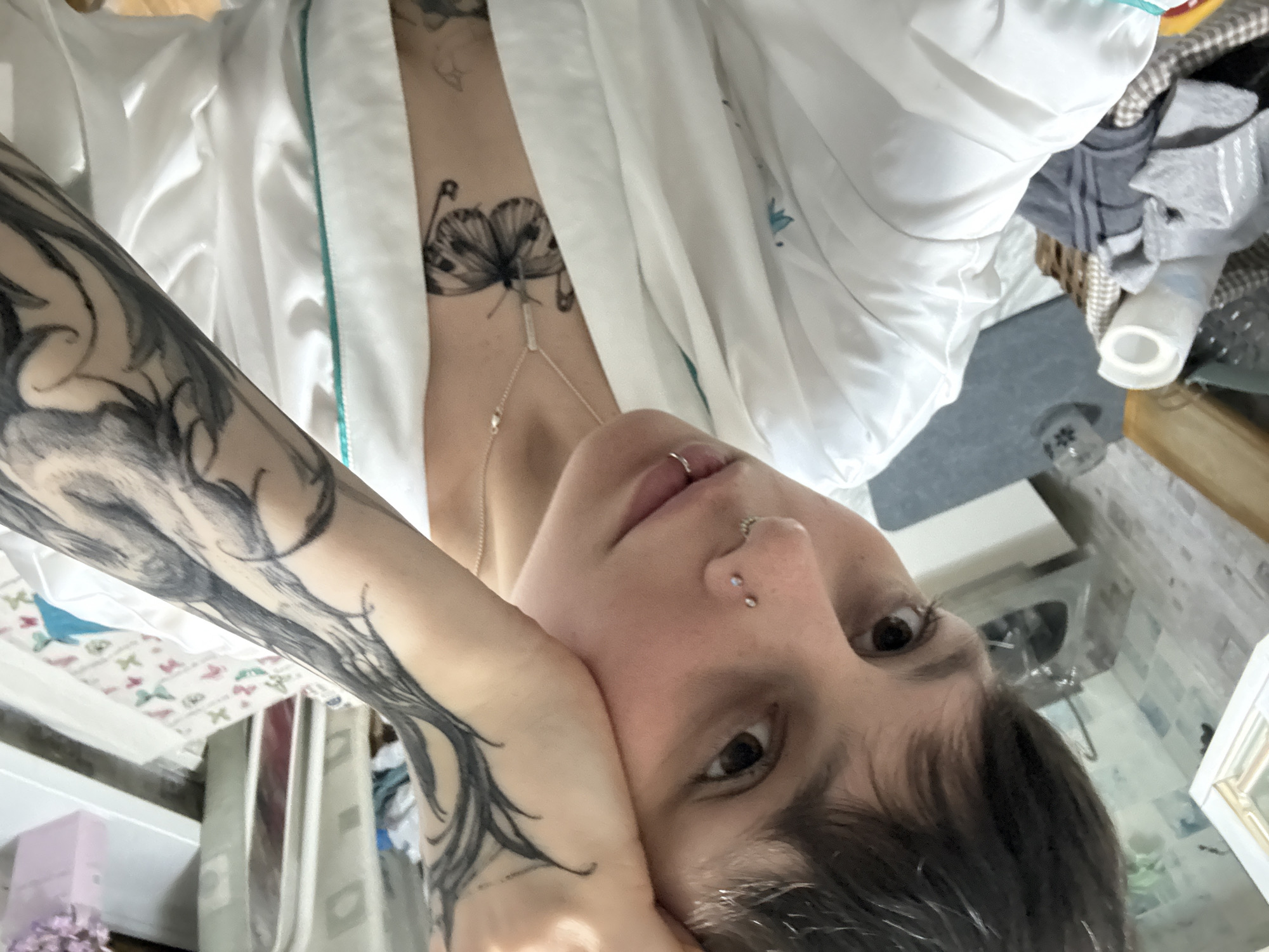 A selfie shows a person with a butterfly tattoo on their upper chest, a tattooed arm, and facial piercings. They are wearing a white robe. The background is blurry and shows parts of a room, including a window with floral patterned fabric, a side table, and what appears to be a cluttered area with assorted items. There is no evidence to confirm a location in the city of Saint Petersburg, Russia.