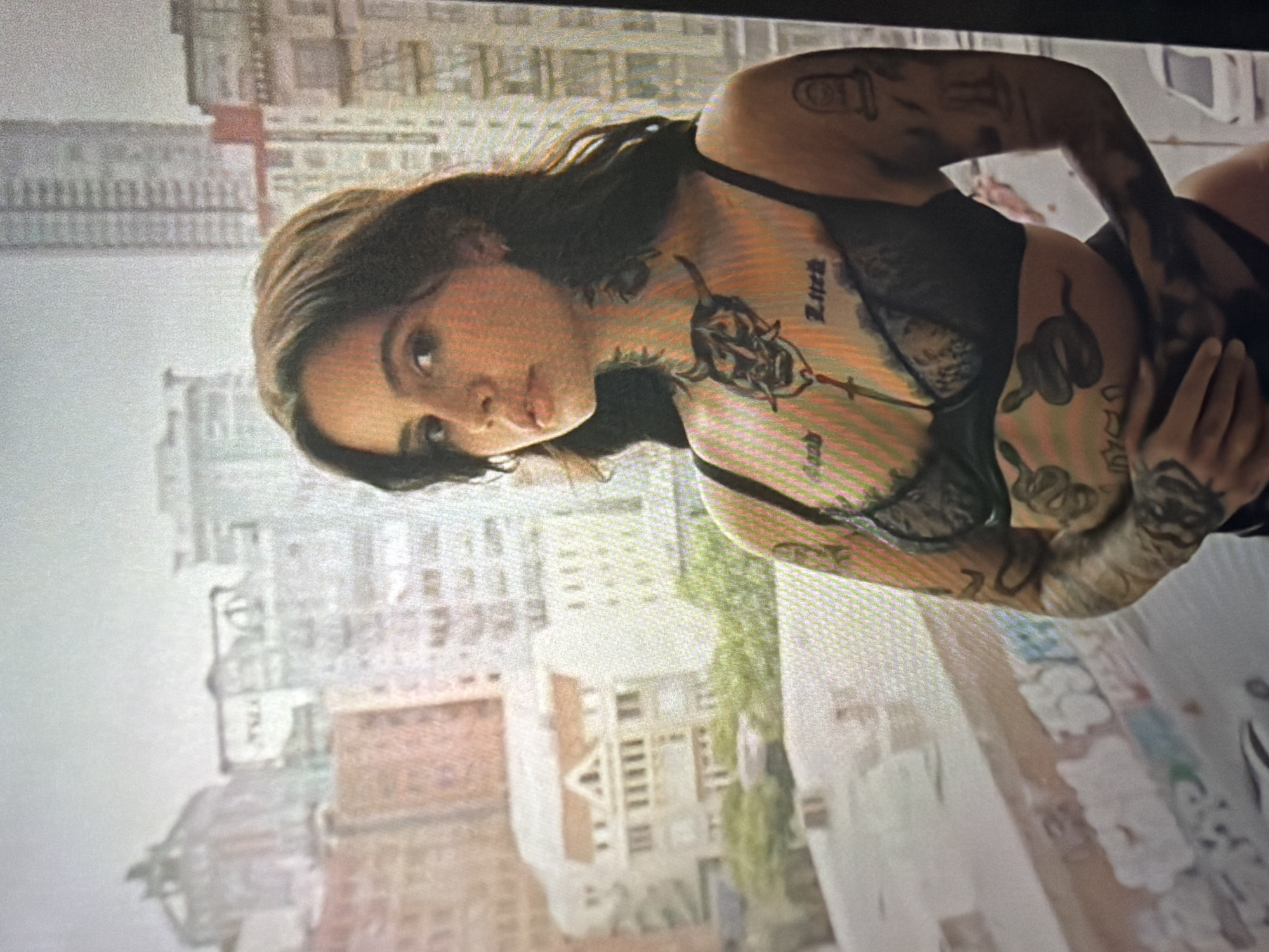 The image shows a woman standing in front of a window with a cityscape in the background. She is wearing a black bra and underwear. She has several tattoos on her body, including a devil mask on her chest, snakes on her stomach, and other designs on her arms and neck. The cityscape appears to be densely populated with various buildings. There is no indication of the location being Shumen, Bulgaria.