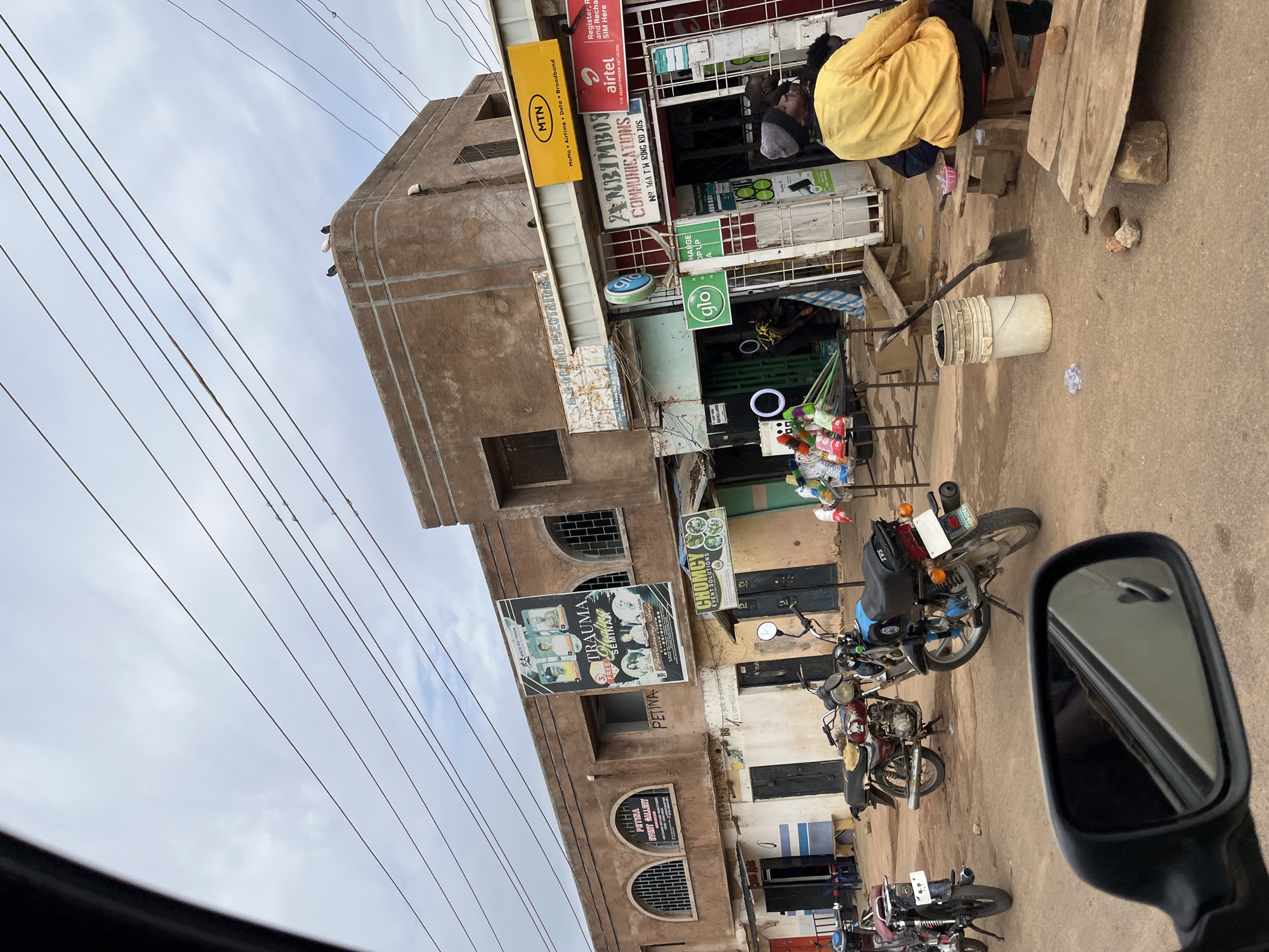 The image shows a street in Jos, Nigeria, with a multi-story building on the left side. The building has a variety of businesses and signage. Noteworthy brands such as MTN, Airtel, and GLO are displayed. A sign for "Zanbimboy Communications" is visible. Other signs advertise "Trauma Seminar" and "Chomcyc Event Solutions". The building is aged with visible weathering.
On the street level, there are motorcycles parked, and a person wearing a yellow jacket sits near the edge of the frame. There is a white bucket and various items displayed, possibly for sale. Overhead power lines crisscross the sky. The view is from the perspective of someone likely in a vehicle, as a side mirror is visible in the lower right corner of the frame.