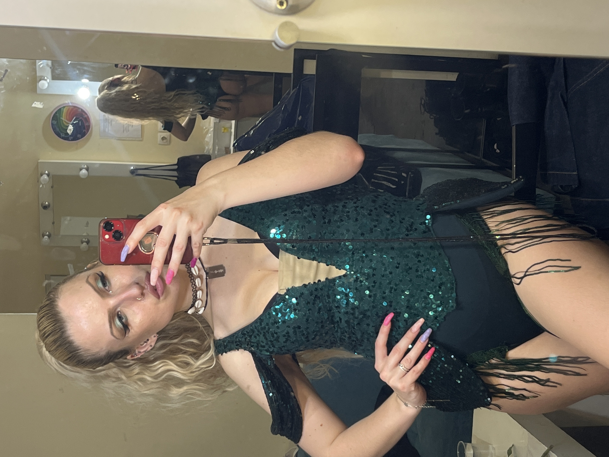 The image shows a person in a dressing room, holding a red smartphone in front of a mirror, taking a selfie. The person is wearing a green sequined outfit with black fringe, and a seashell choker with a razor blade charm. They have long blonde hair and colorful nail polish. The dressing room contains a makeup mirror with lights, a clothes rack, and various items on shelves. A second person can be seen reflected in the mirror, wearing a black outfit. While the image provides no explicit evidence of the location being in Plovdiv, Bulgaria, it suggests an indoor dressing room environment associated with performance or costume activities.