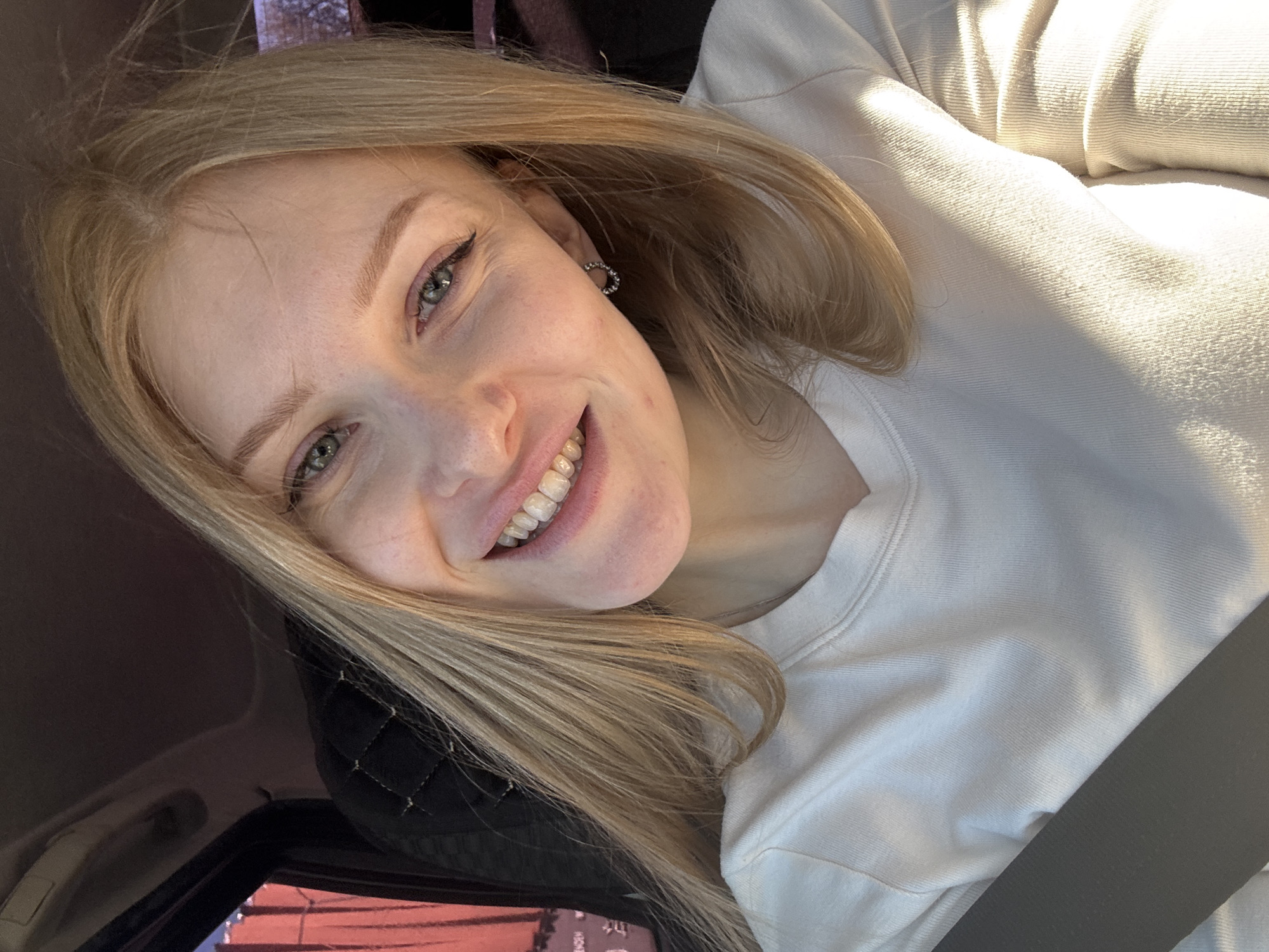 A young adult female with light blonde hair and fair skin is in the driver's seat of a vehicle. She is wearing a white long-sleeved shirt and has a seatbelt fastened across her chest. Her hair is parted and falls loosely around her face. She is smiling, exposing her upper teeth. She is wearing small, circular earrings. The interior of the vehicle is visible, including the dark upholstery of the seat and a glimpse of the car's dashboard or interior trim. The location of the image is likely within a vehicle and is not identifiable as being in Perm, Russia based on the image content. The exact geographical location cannot be determined from the information provided.