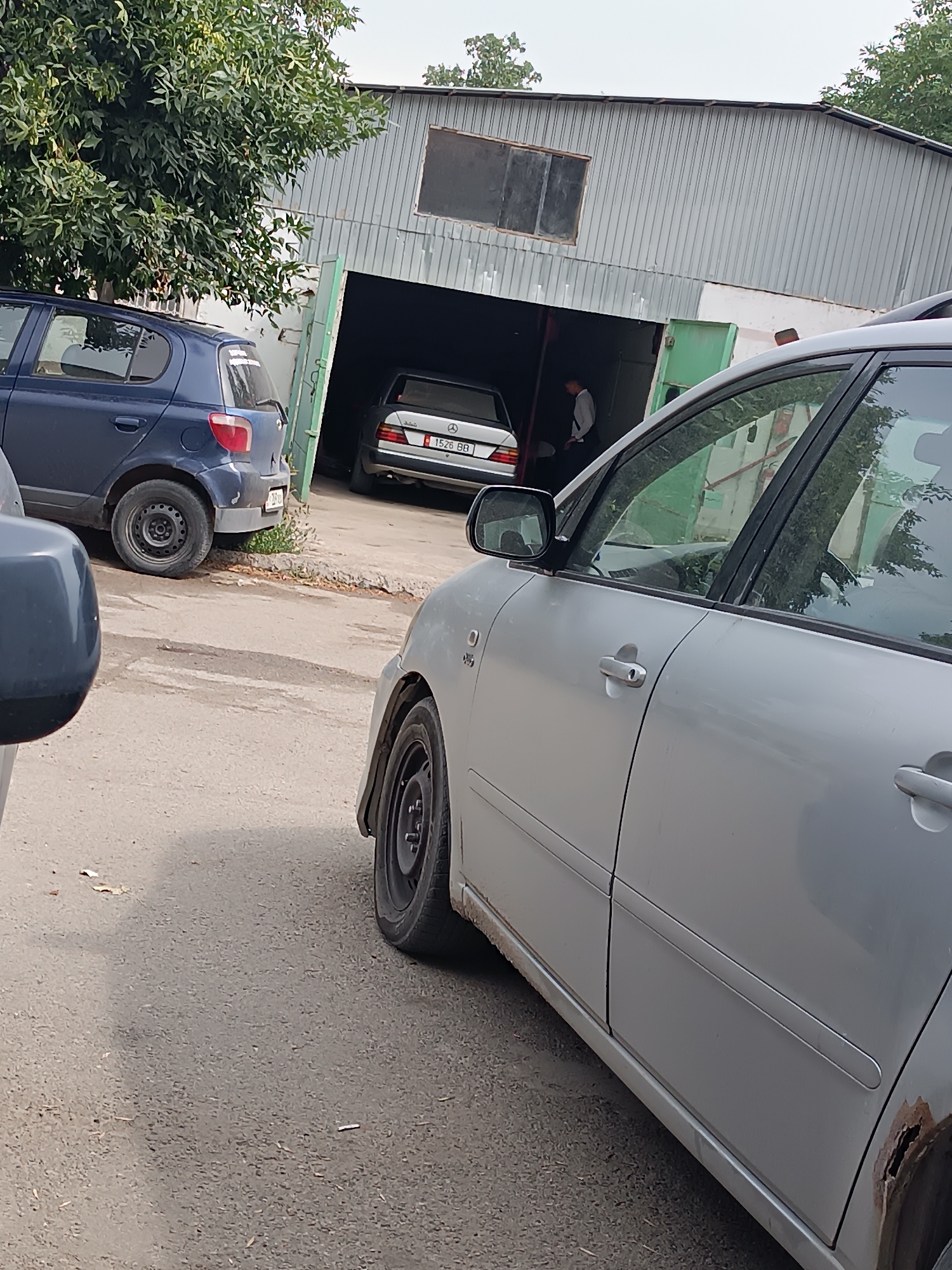The image shows a view of a garage and its surroundings. In the foreground, a silver car is parked. A dark blue car is parked to the left of the garage. Inside the garage, a silver Mercedes-Benz is visible. A person in a white shirt and dark pants is standing in the garage. The garage structure appears to be made of corrugated metal. There are trees and vegetation in the background. The ground is paved.