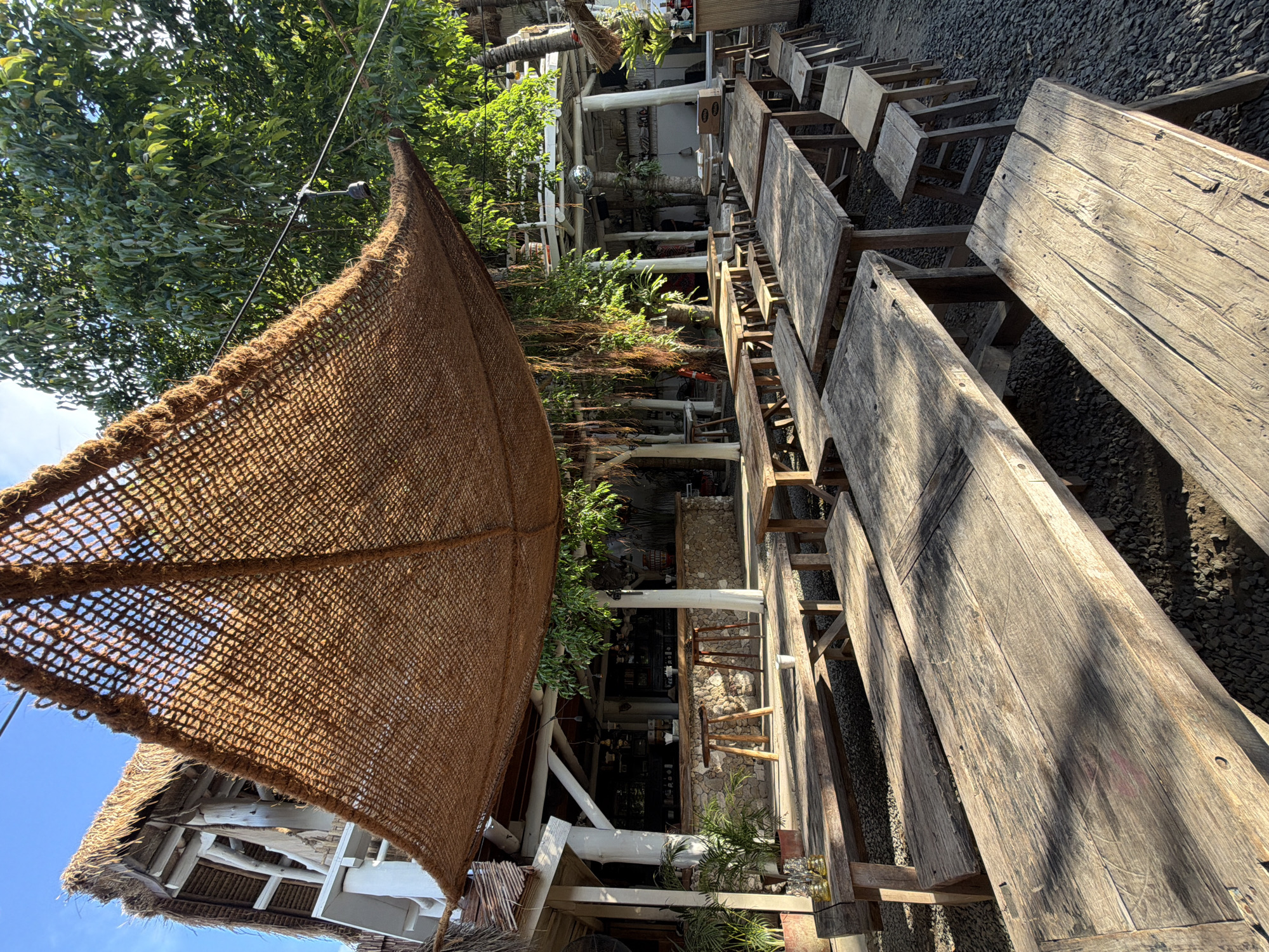 The image shows an outdoor seating area with rustic wooden tables and benches, set against a backdrop of lush greenery and a building with a thatched roof. The scene appears to be a cafe or restaurant in Kute, Indonesia, with a laid-back, tropical ambiance.
The seating area is sparsely furnished with simple, weathered wooden tables and benches, arranged on a gravel-covered ground. Some tables have chairs or stools around them, while others are set up as picnic-style seating. A large, brown woven canopy provides shade over some of the tables.
In the background, a two-story building with a thatched roof and white walls is visible. The building has an open-air design, with exposed beams and balconies. A bar area with stone accents is visible inside the building.
Plants and trees are abundant, with tropical foliage providing a natural and refreshing setting. Hanging vines and potted plants add to the tropical feel.
The image captures a serene and relaxed atmosphere, suggesting a place where people can unwind and enjoy the tropical surroundings. The overall impression is one of a charming and unpretentious outdoor dining or gathering space.