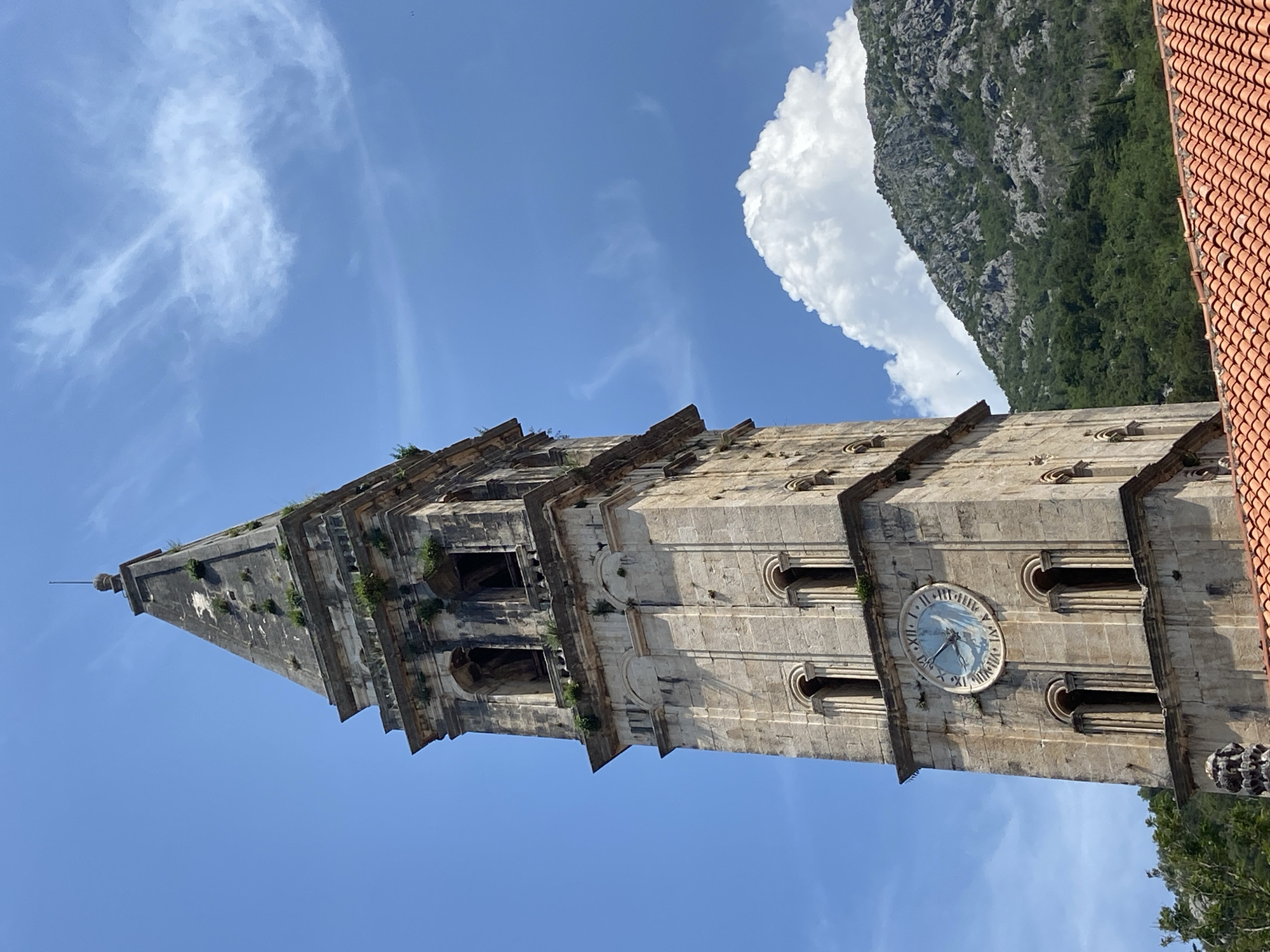 This is a low-angle, medium shot of a tall, stone bell tower in Perast, Montenegro. The tower has a clock face with Roman numerals. Above the clock are arched windows. The tower is topped with a spire. The sky is blue with white clouds. In the background, a green, rocky mountain is visible. In the foreground, red tile roofs of buildings are present.