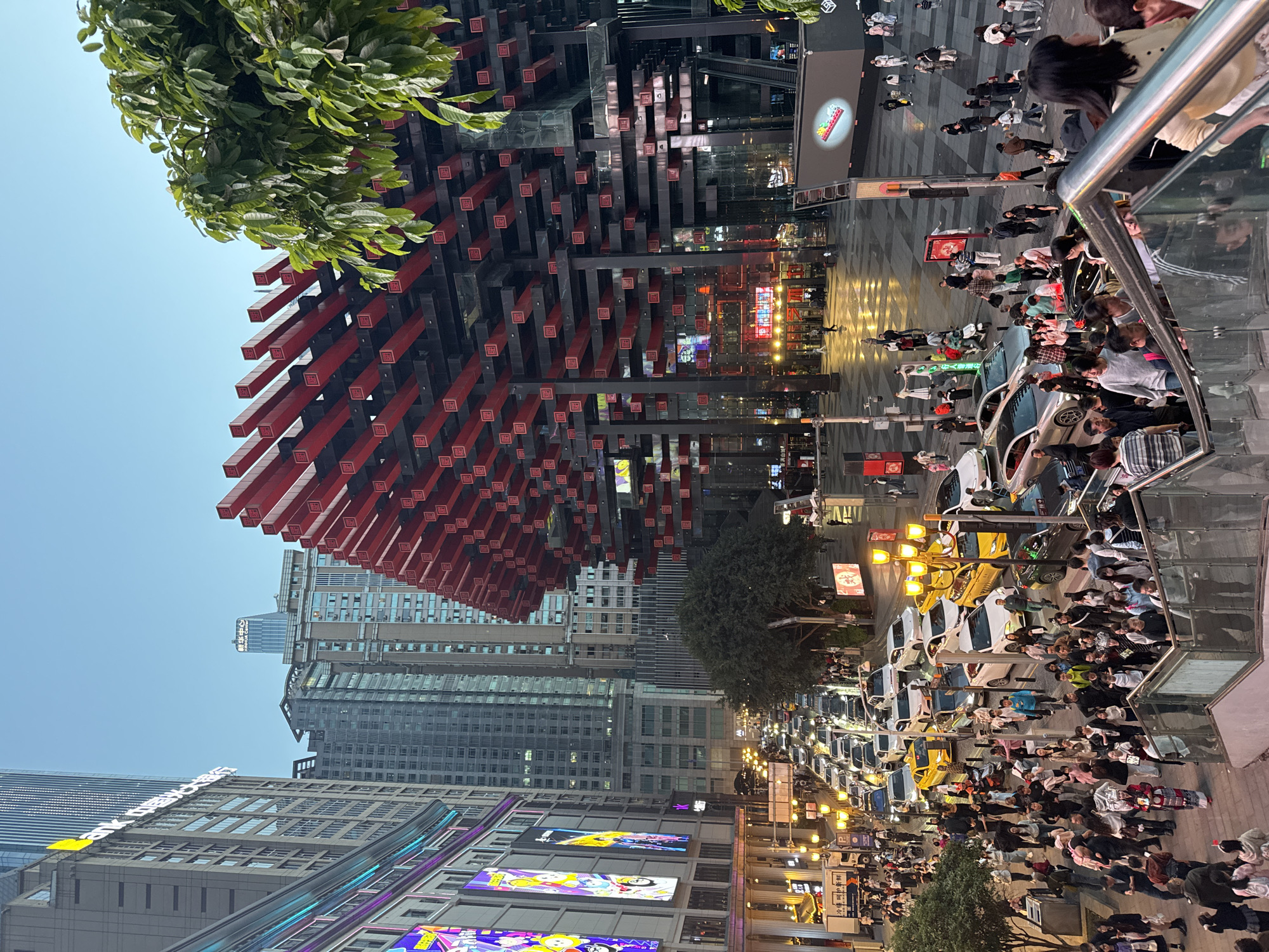 The image depicts a bustling urban scene in Yuzhong District, Chongqing, China. A distinctive architectural structure composed of numerous red and black columns dominates the upper-left portion of the frame, contrasting with the surrounding modern skyscrapers. A dense crowd of people fills the street below, intermingling with vehicles, including many taxis, creating a vibrant street-level atmosphere. Illuminated billboards and streetlights add to the urban vibrancy, particularly noticeable during the evening. The image captures the dynamic interplay of architecture, pedestrian activity, and urban infrastructure in a busy commercial area of the city.