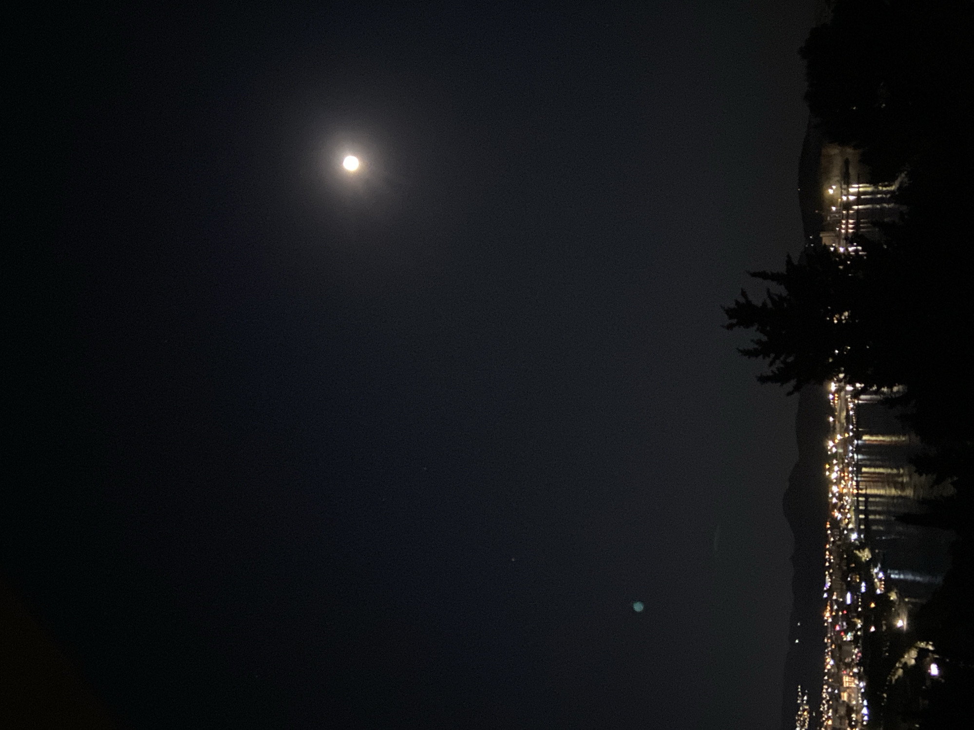 The image shows a nighttime view in Šušanj, Montenegro. The night sky is dark with a bright moon. Below the horizon, there is a dense concentration of lights, suggesting a city or town along the coastline. The lights reflect on the water. Silhouettes of trees are visible on the right side of the image.