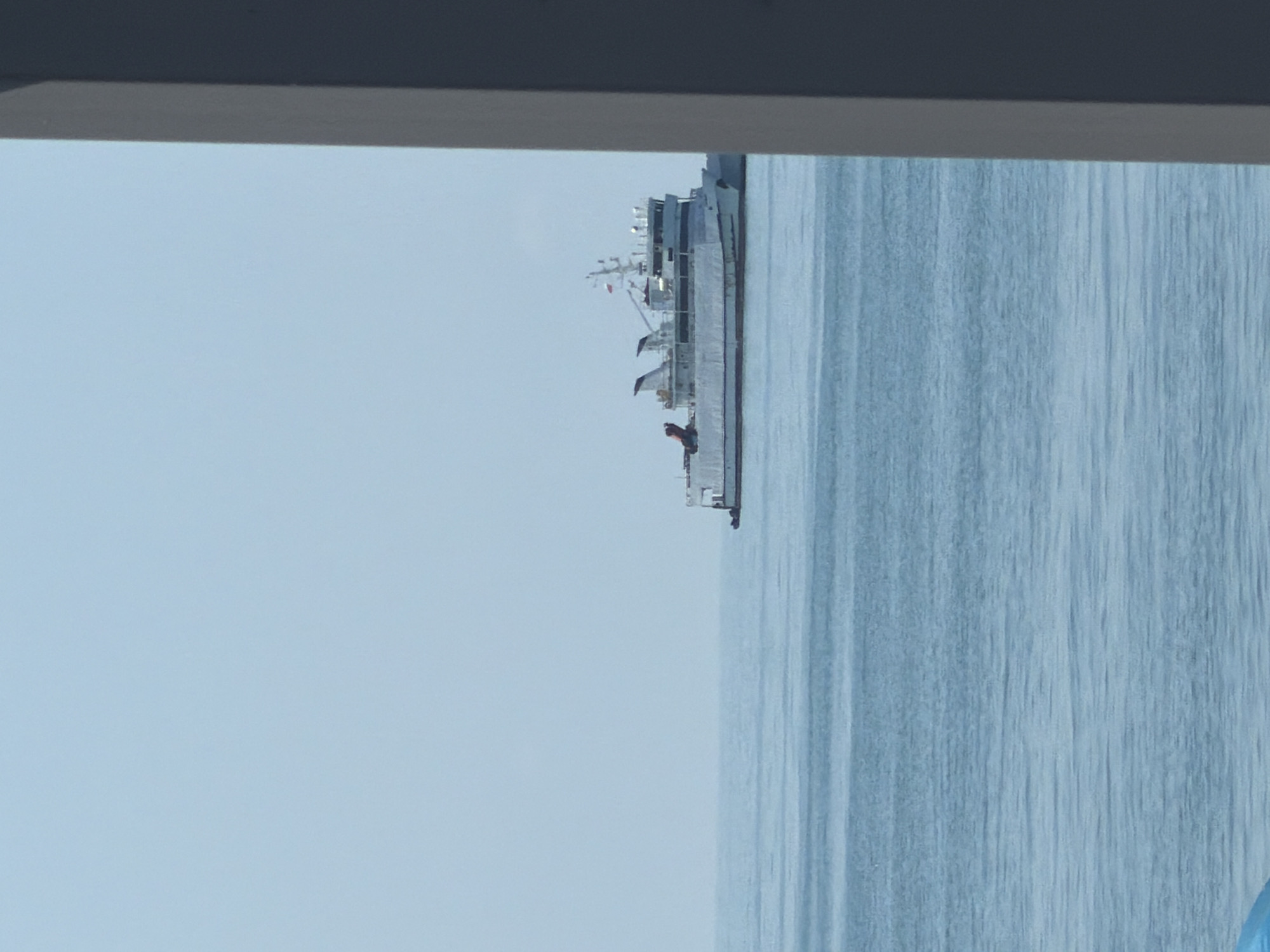 A white ferry boat with a red and blue stripe on its side is sailing on a calm blue sea under a clear blue sky. The boat has multiple decks and a bridge. The scene is partially framed by a dark gray vertical structure on the right side of the image. The water is flat with subtle ripples. The location is the city of Sochi, Russia.