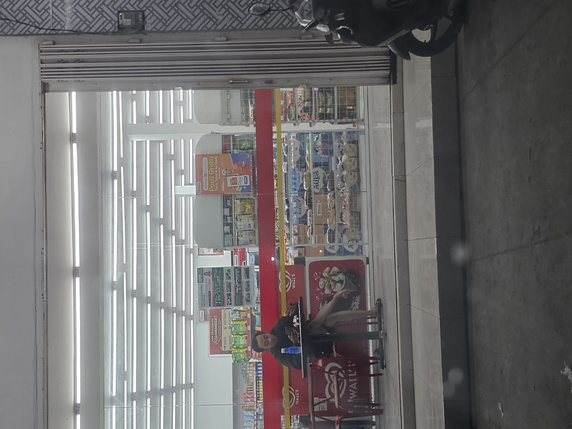 A man is sitting at a small, black, round table outside a convenience store in Sumberwudi, Indonesia. He is wearing a dark-colored jacket and dark-colored shorts, with his legs crossed. He is holding a smartphone in his left hand and appears to be eating. A blue bottle of water is on the table, as well as a small plate with food. There are two red chairs at the table, but only the one the man is sitting on is visible. The store has a red and white facade and is brightly lit from within by fluorescent lights. A large advertisement featuring ice cream is visible on the store's exterior. A scooter is parked to the right of the entrance. The pavement in front of the store is grey.