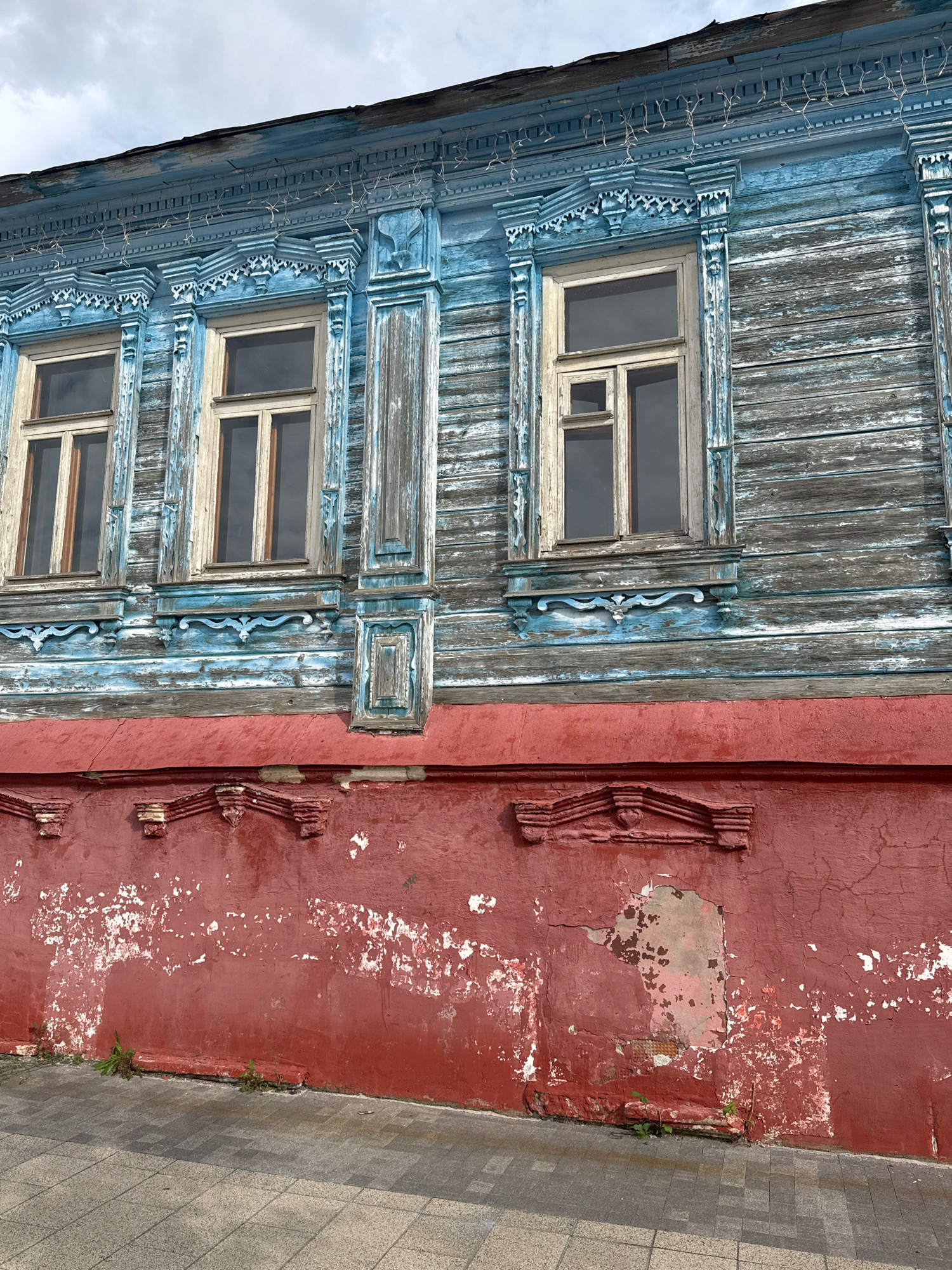 The image shows a close-up view of the facade of an old wooden house in Zaraysk, Russia. The upper part of the house is painted a faded blue, with weathered horizontal wooden planks. Intricate wooden carvings adorn the eaves and window frames. There are two windows visible, with light-colored frames and multiple panes. A string of small white lights is draped along the top of the facade. The lower part of the house is a red-painted wall, which shows significant signs of wear and peeling paint, revealing patches of a lighter color underneath. Decorative elements are carved into the red wall as well. A concrete sidewalk with a patterned surface runs along the base of the wall. Small green weeds are growing near the base of the red wall. The sky above is overcast.