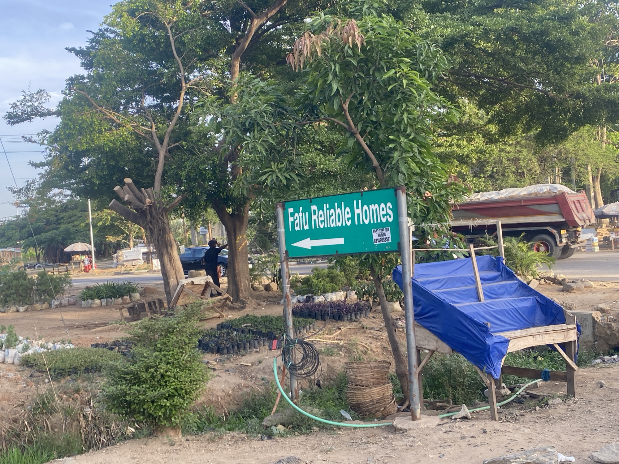 The image depicts a roadside scene in Karmajiji, Nigeria. A green sign reading "Fafu Reliable Homes" with a left-pointing arrow is prominently displayed. To the right of the sign is a wooden structure covered in blue tarpaulin. In the background, a red truck is parked on the side of a road. Across the road are several trees and a person dressed in black leans against a tree. Numerous potted plants are arranged on the ground in front of the trees. A coiled hose and a woven basket are near the signpost. The ground is dirt.