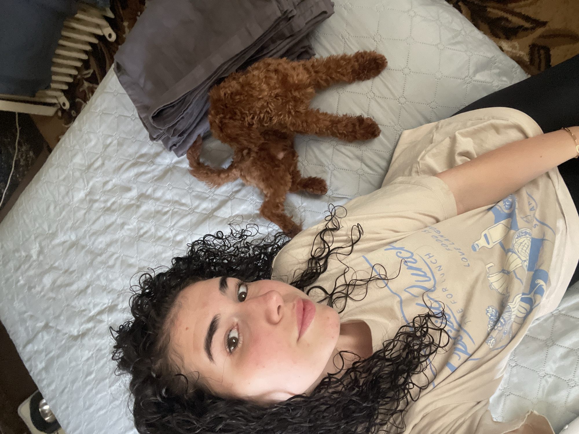 A woman with curly black hair, lying on a light blue quilted bedspread, takes a selfie. Her head is at the bottom left of the frame. She is wearing a beige T-shirt with light blue writing and illustrations. A reddish-brown poodle is lying head-down near the top center of the frame, next to a pile of gray fabric. Part of a white radiator is visible in the upper left corner. The location cannot be determined based solely on visual information in the photo.