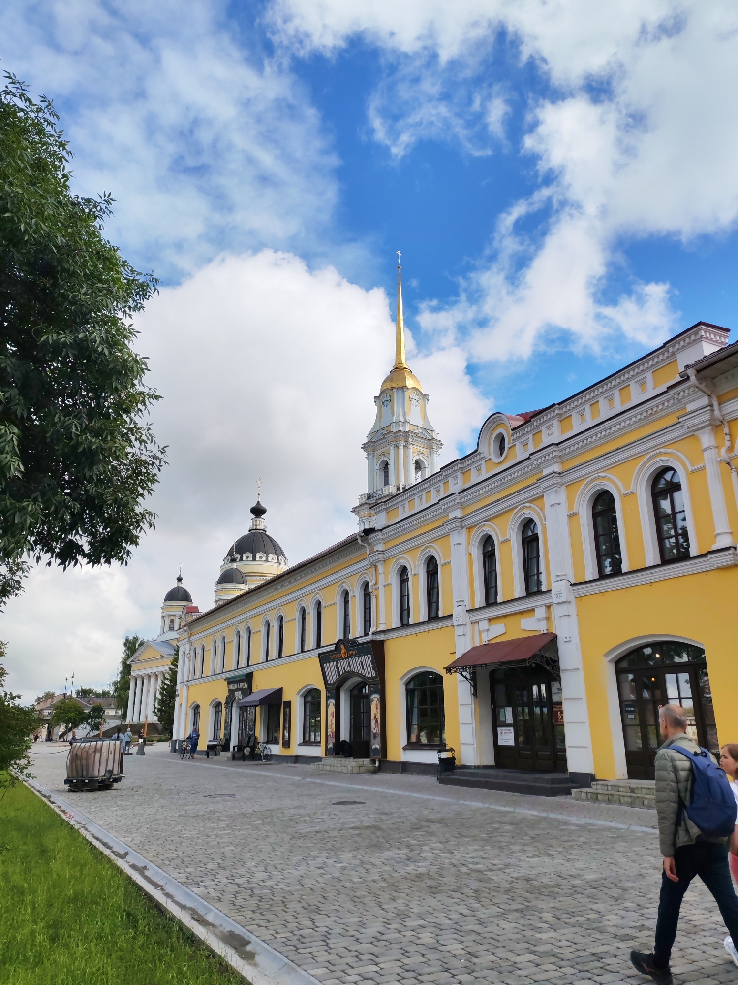 In Bol'shie Mkhi, Russia, the image captures a street scene with a prominent yellow building with white architectural details. The building features arched windows and a gold-tipped steeple that towers against a blue sky with scattered clouds. A dark-domed building with a cross is visible behind the yellow structure. In the foreground, a cobblestone street is lined with green grass on the left side. A large, cylindrical wooden object sits on the street near the grass. Two people, one with a blue backpack, are walking away from the camera on the right side of the street. The scene appears to be a public area, possibly a town square or a street in a historic district.