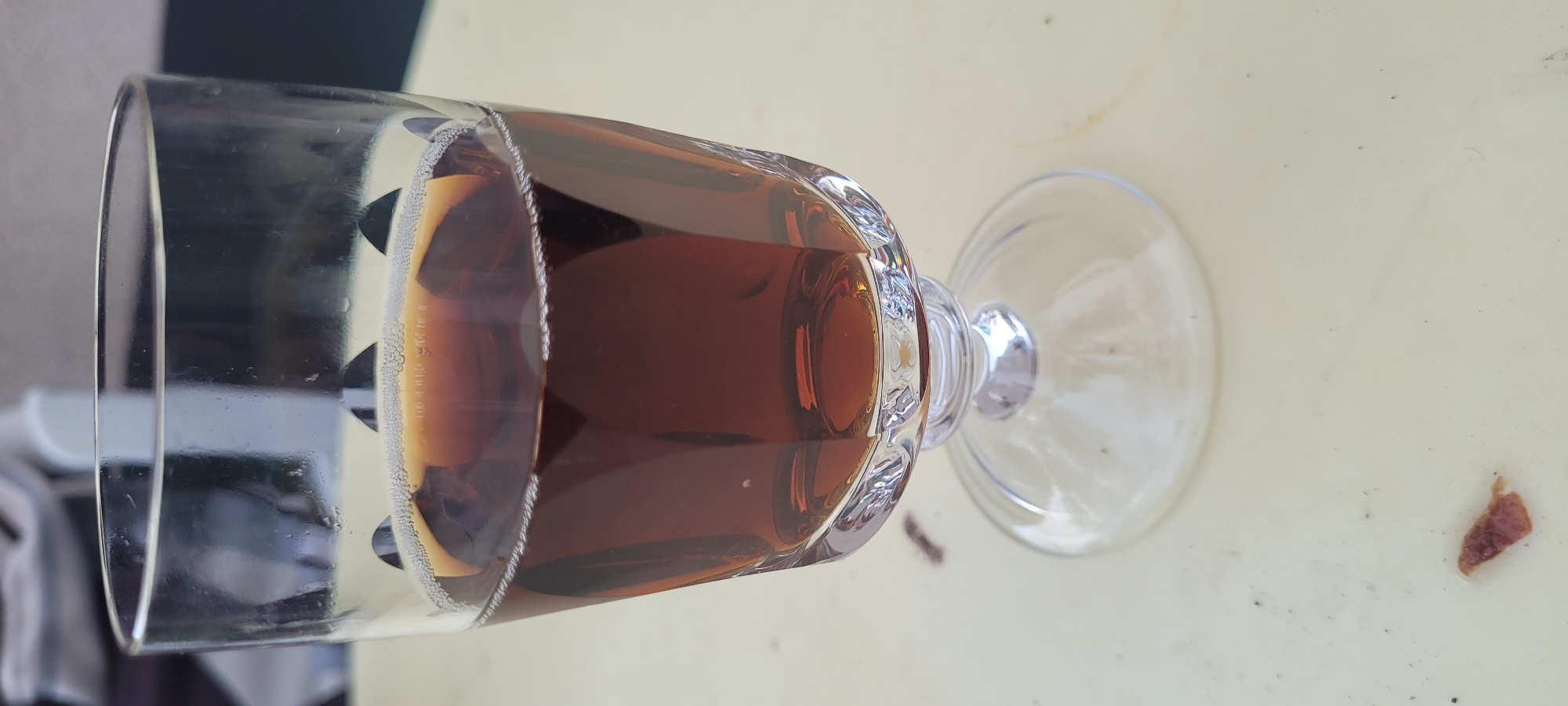 A close-up, eye-level shot shows a stemmed glass filled with a dark amber liquid, possibly iced tea or a similar beverage. The glass has a faceted design and rests on a clear, round base. The liquid has a thin layer of small bubbles near the rim. The glass is positioned on a light-colored, textured surface that appears to be a table. In the blurred background, a person in a chair is partially visible, suggesting an outdoor or semi-outdoor setting. A small, dark object, possibly food debris, is on the table in the foreground. The image does not provide any specific information about the city of Dietzenbach, Germany.
