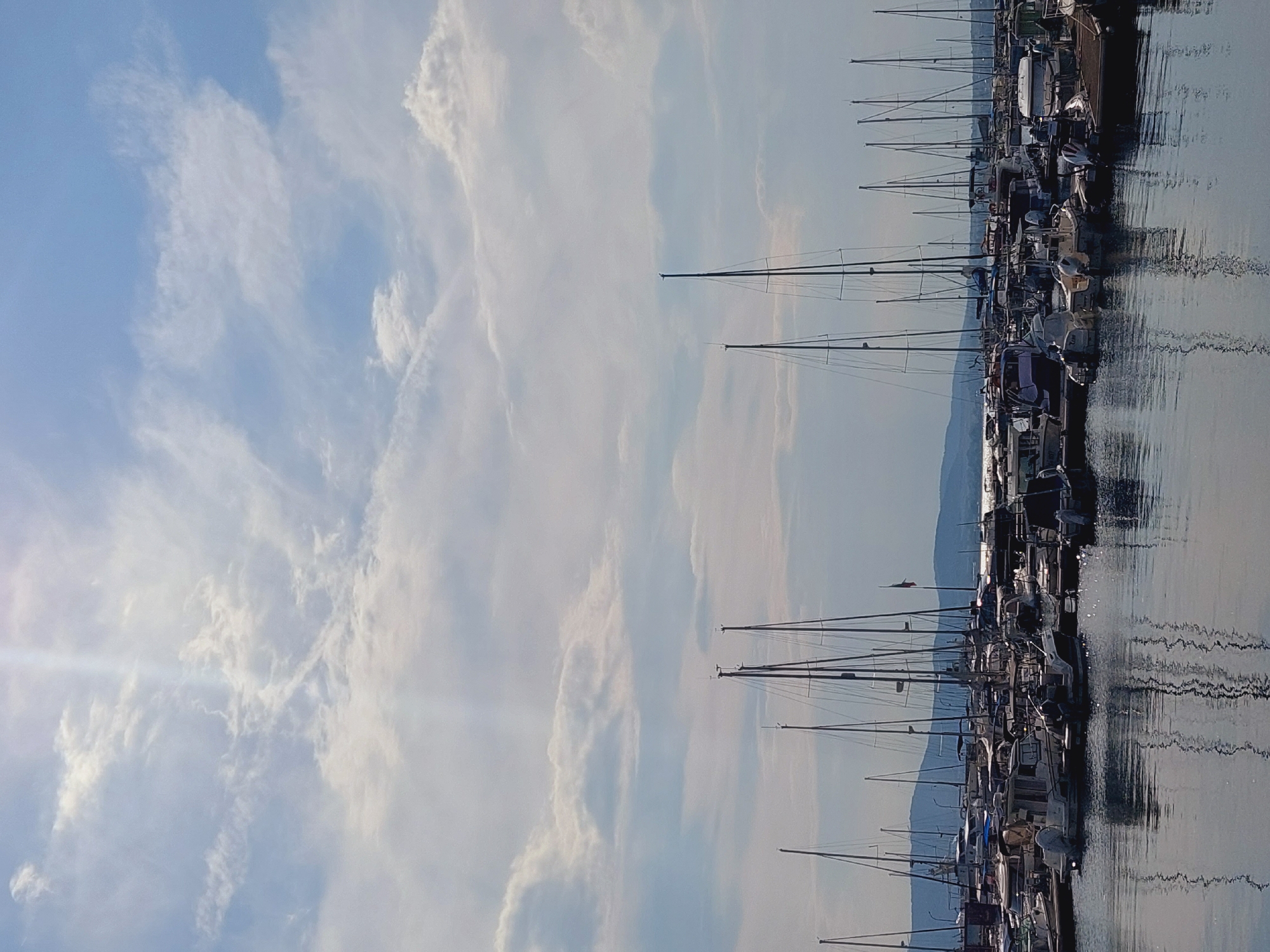 A daytime, eye-level, vertical, medium shot captures a harbor filled with numerous docked sailboats and motorboats. The boats, varying in size and design, are moored in rows and fill the lower half of the frame. Their reflections are visible in the calm, dark water. In the background, a distant shoreline with rolling hills is visible under a partly cloudy sky. The sky is a mix of blue and white, with streaks of sunlight breaking through the clouds, creating a bright, ethereal effect. The scene is characteristic of a maritime area in Sozopol, Bulgaria.