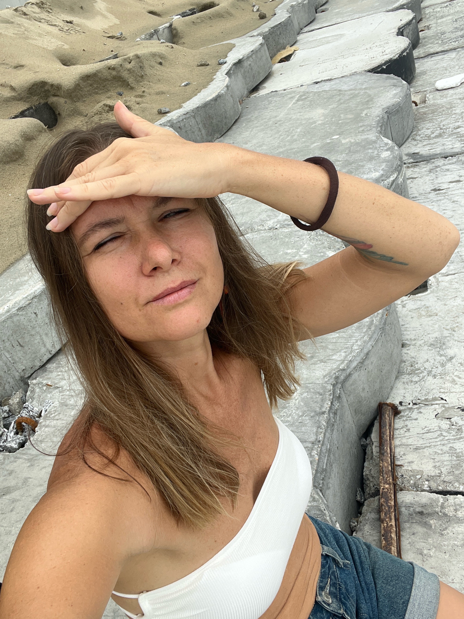 A woman with long, light brown hair shades her eyes with her right hand as she looks towards the camera. She is wearing a white strapless top and a denim jacket. Her left arm is bent, and a dark brown hair tie is visible on her wrist. A tattoo is visible on her upper left arm. She is sitting on concrete structures, with sand in the background. The scene appears to be outdoors, possibly near a beach or coastal area. The image is a close-up, with the woman's face and upper body filling most of the frame. There is no visual information in the image that would indicate the location is Hoi An, Vietnam.