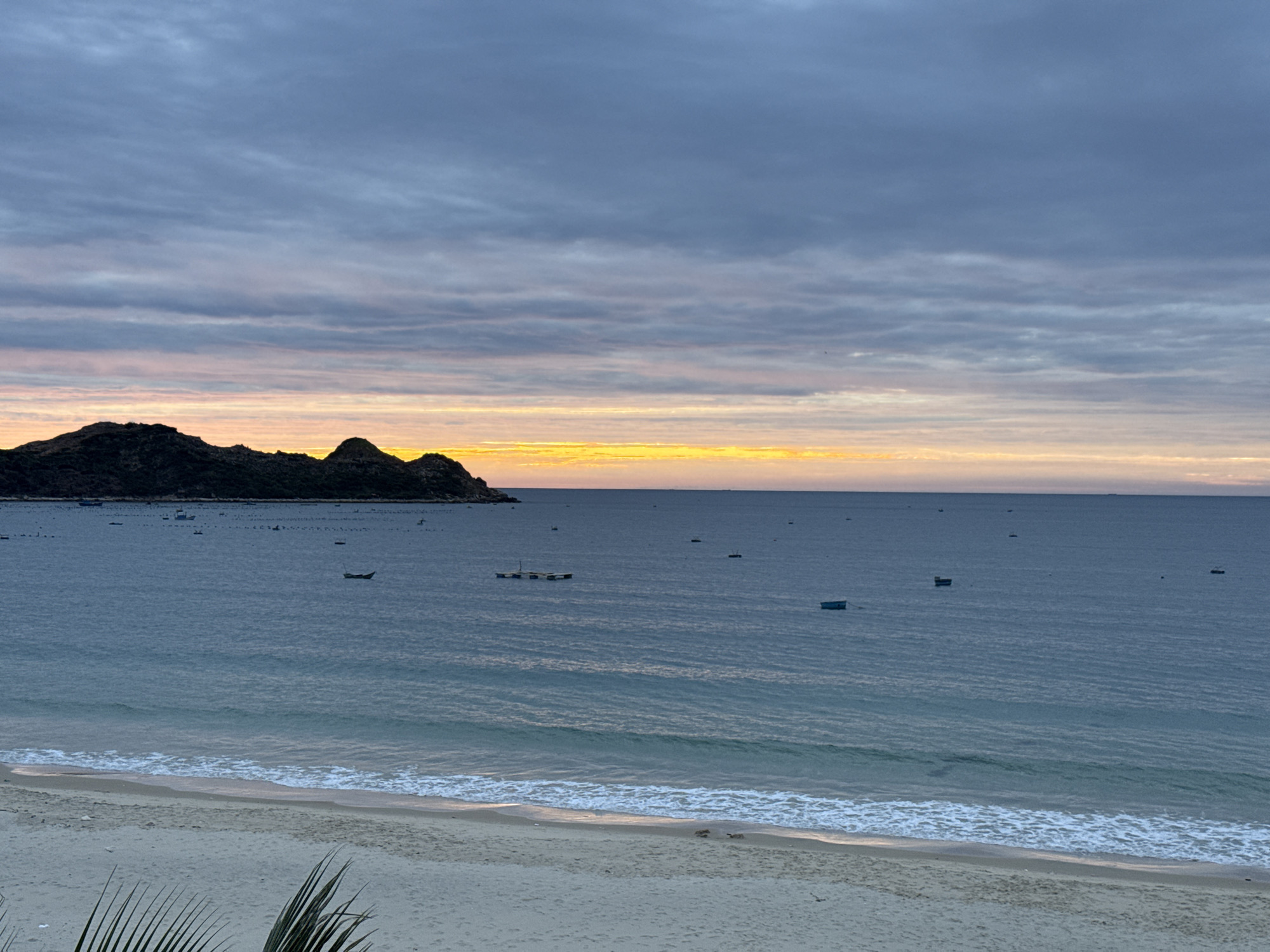 The image depicts a serene coastal scene at dusk in Song Cau, Vietnam. The foreground shows a sandy beach with gentle waves lapping at the shore. Beyond the beach, the calm ocean stretches towards the horizon, dotted with several small boats. In the distance, a rugged, dark landmass with a few structures is visible, possibly representing the coastline of Song Cau. The sky above is a canvas of muted colors, with streaks of orange and pink from the setting sun blending into shades of blue and gray clouds. The overall impression is one of tranquility and natural beauty.