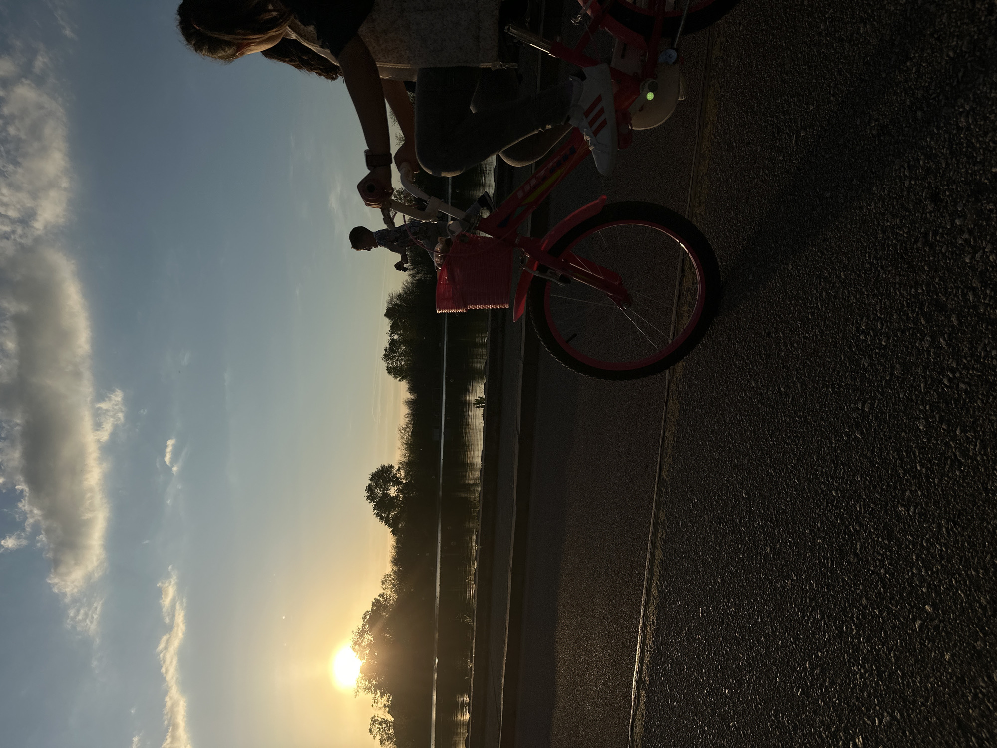 A low-angle, horizontally-oriented shot depicts two people riding bicycles, probably a mother and child, in Plovdiv, Bulgaria, at dusk. The child rides a red bike with a pink basket attached to the front. The woman, partially visible at the top, wears sneakers with three stripes. The sunset casts a glare over a body of water, with trees silhouetted along its edge, and a sky with clouds. A section of pavement occupies the bottom right.