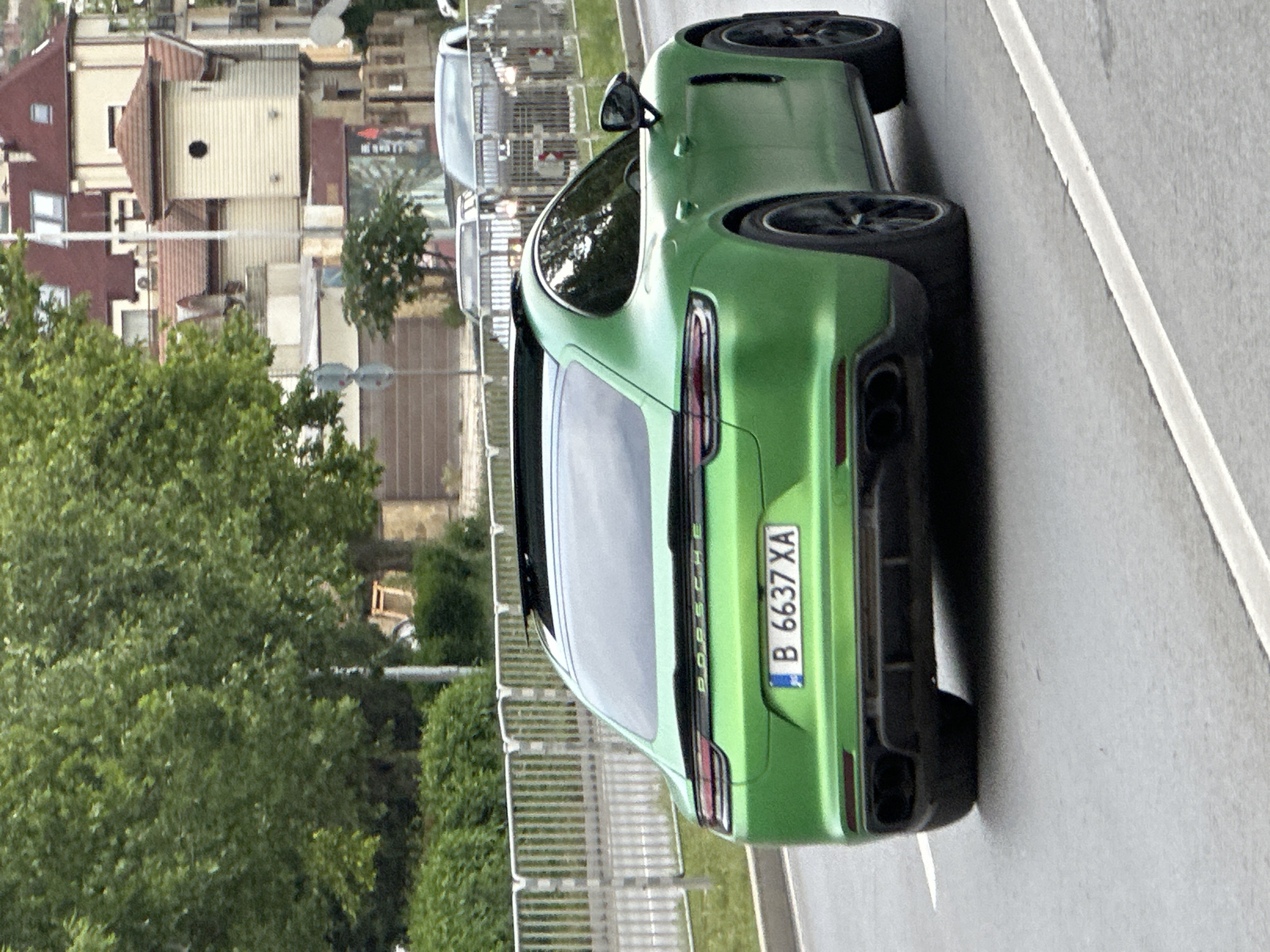 A vibrant green Porsche Panamera Sport Turismo is the central subject of this daytime image, captured from a slightly elevated rear-quarter angle. The car features black multi-spoke wheels, a sporty rear diffuser, and prominent dual exhaust tips. "PORSCHE" is emblazoned across the rear tailgate, below the tinted window. Its license plate, clearly visible, reads "B 6637 XA," with the 'B' indicating registration in Sofia, Bulgaria.
The car is situated on the right side of an asphalt road, which has a distinct solid white lane marking. In the background, a residential area unfolds with various buildings, some featuring reddish-brown tiled roofs and light-colored facades. Abundant green trees and shrubs line the street, separated from the roadway by a metal fence. Another, lighter-colored vehicle is partially visible further down the road, behind the Porsche. The lighting suggests clear, bright weather conditions during the day. The image itself is oriented vertically, appearing rotated 90 degrees counter-clockwise.