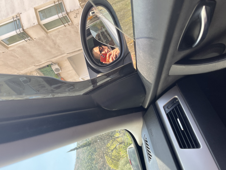 The image depicts a scene captured from the interior of a vehicle, presenting a view outwards into an urban environment in Budva, Montenegro, under bright daylight.
**People:**
One individual is visible in the passenger-side exterior mirror reflection. This person is holding a red smartphone, positioned as if taking a selfie, and wears dark-framed glasses. Only the upper facial area and part of the shoulders are discernible.
**Subjects/Objects:**
The car's interior is partially visible in the foreground, showing a dashboard with black and silver accents, an air vent, and an "AIRBAG" label. The dark grey A-pillar and a section of the door panel with a chrome handle are also present. Through the front passenger window, to the right, a multi-story building with a light-colored, possibly stucco, facade is visible. It features several windows, some with external railings, and an externally mounted air conditioning unit. To the left, a natural, green hillside covered with trees, including a prominent slender conifer, rises against a clear blue sky. A dark-colored vehicle is partially visible in the lower-left, obscured by the car's interior.
**Actions/Interactions:**
The visible individual is engaged in self-photography, using the car's exterior mirror to compose the shot. The perspective suggests the car is either stationary or moving slowly along a street bordered by buildings and natural terrain.
**Environment:**
The environment combines urban architecture with natural hilly landscapes, indicative of Budva's geographical characteristics. The lighting is strong and consistent with a clear, sunny day.