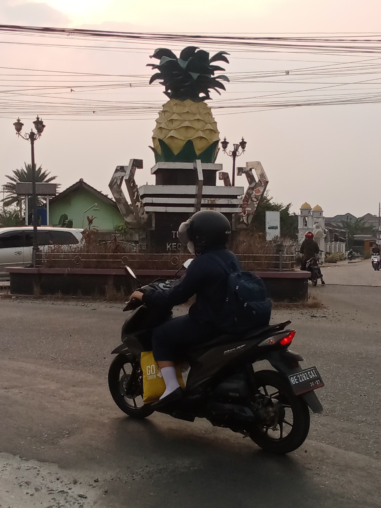 A rear-view, medium shot captures a person riding a black motorcycle on a paved road in what appears to be Toto Katon, Indonesia, as indicated by the text on a nearby monument. The rider, wearing a black helmet and a dark blue jacket over a white shirt, carries a blue backpack. A yellow bag with the words "GO GREEN" printed on it is placed on the motorcycle's footrest. The motorcycle's license plate reads "BE 2282 GAI".
In the background, a large pineapple-shaped monument stands prominently, adorned with decorative structures and streetlights. Behind the monument, a modest green building is visible, along with trees and other structures. Further in the distance, two minaret-like structures with yellow domes suggest the presence of a religious building. The sky is overcast with subtle hues of pink and orange, indicating it might be dusk or dawn. The road surface shows signs of wear and tear, with scattered gravel. Other motorcycles are also seen in the distance, suggesting traffic.