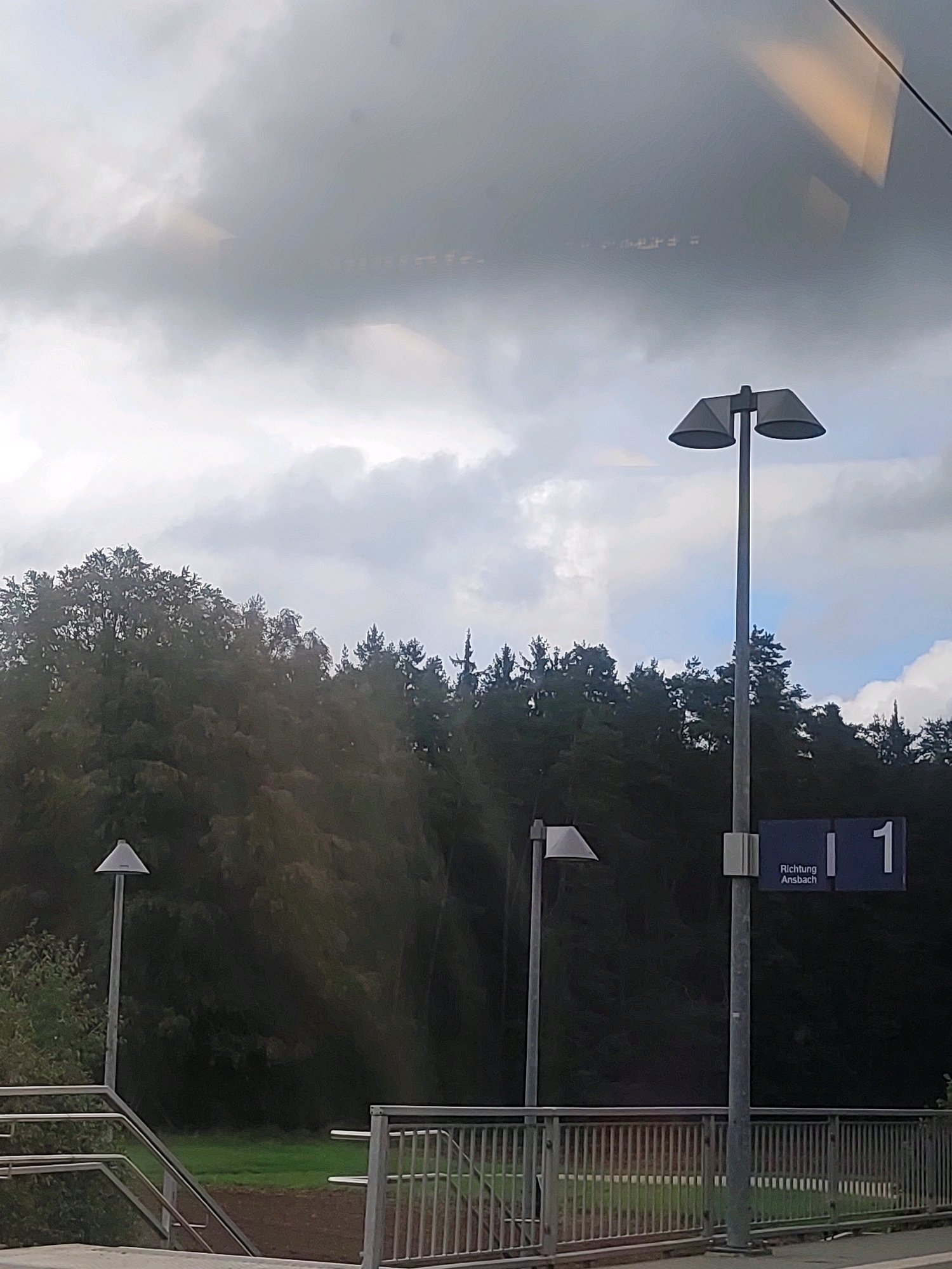 The image shows a train station platform in Petersaurach, Germany. A lamppost stands in the foreground, to the right of center, with two downward-facing lights. Attached to the lamppost is a sign with the text "Richtung Ansbach" and the number "1". A second, smaller lamppost is visible to the left. In the background, a dense forest with dark green foliage lines the edge of the platform. The sky above is filled with a mix of gray clouds and patches of blue. Metal railings and stairs are also visible, indicating a pedestrian area or access point to the platform. The scene appears to be captured during daylight.