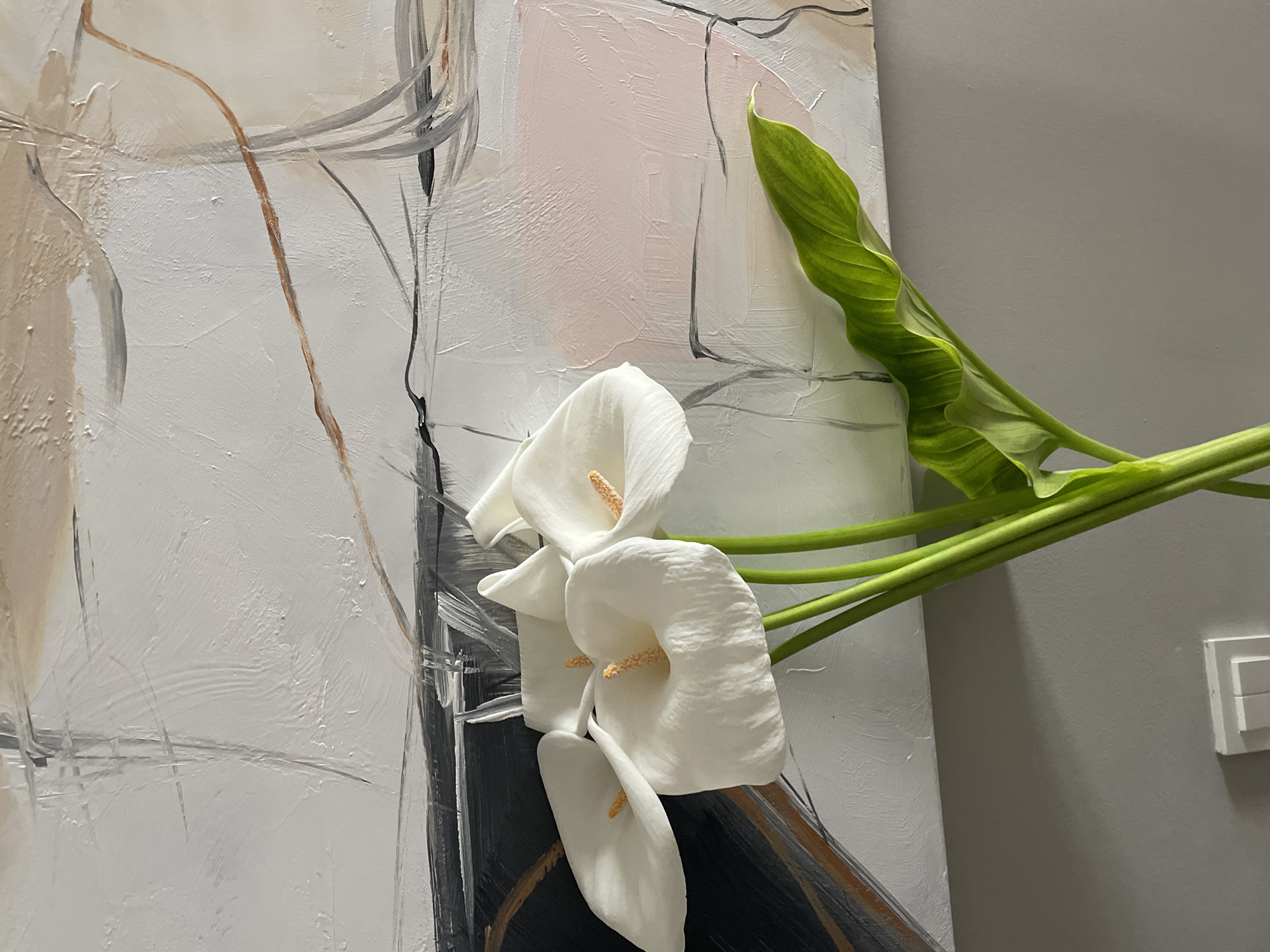 The image depicts a still life arrangement. In the foreground are three white calla lilies with yellow pistils, along with a curved green leaf, arranged against a backdrop. The backdrop is a contemporary painting with abstract strokes of white, gray, black, and a touch of pink. The painting features textured surfaces and appears to be oil or acrylic on canvas. The setting includes a neutral gray wall to the right, where a light switch is mounted. While the image does not specify the location in Tbilisi, Georgia, it indicates a setting that is likely a contemporary interior, possibly a home, studio, or gallery space. There are no people visible in the image.