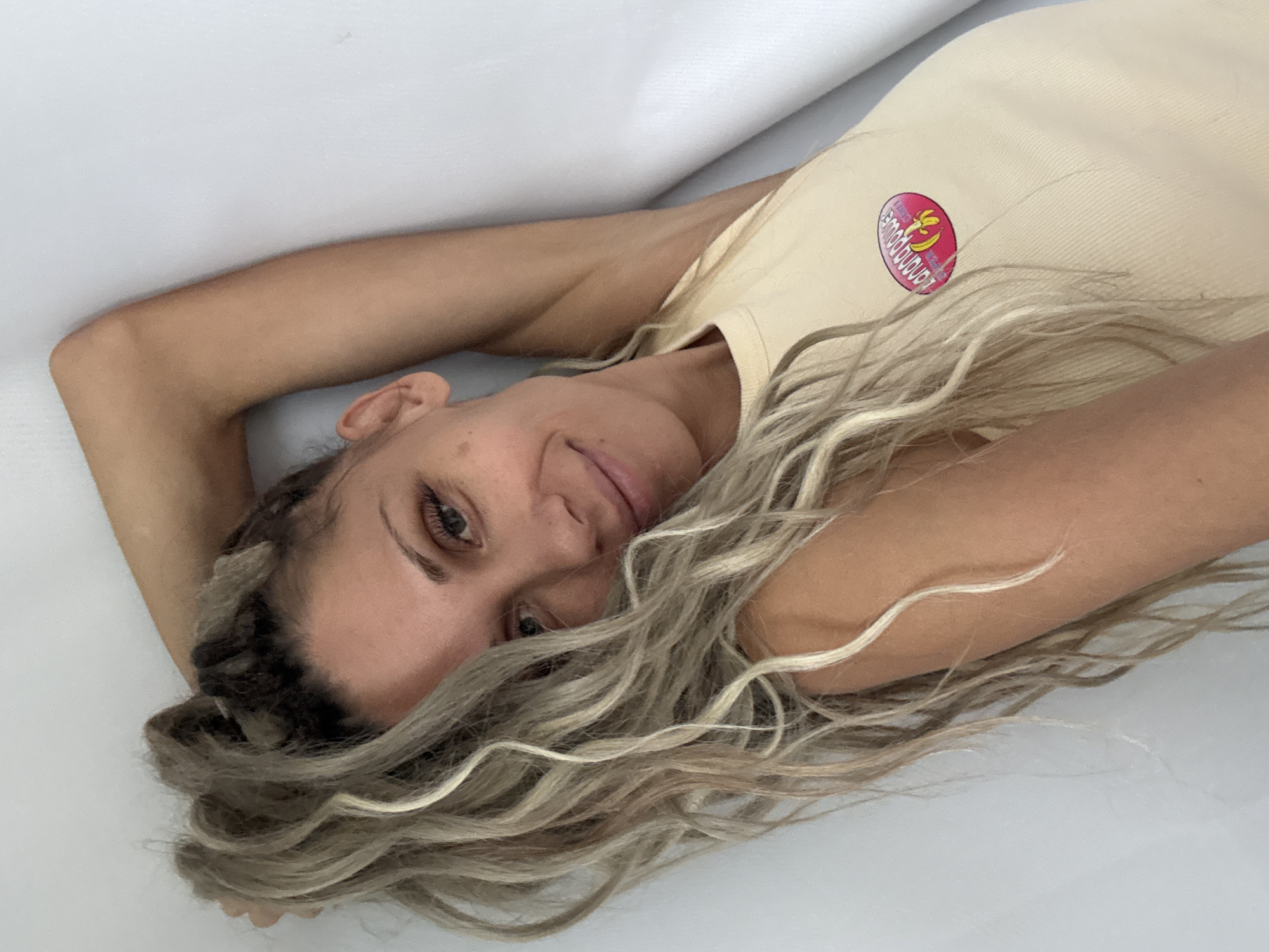 A close-up, waist-up shot shows a woman leaning against a white wall with her right arm raised behind her head. She has blonde hair with lighter streaks, styled in a high ponytail. Her gaze is directed towards the camera, and she has a slight smile. She is wearing a light yellow tank top with a pink and yellow sticker on the left side of her chest that appears to depict a banana and the words "BANANA POWER". The background is a plain white wall. No specific location in Rusanovo, Russia can be determined from the image.
