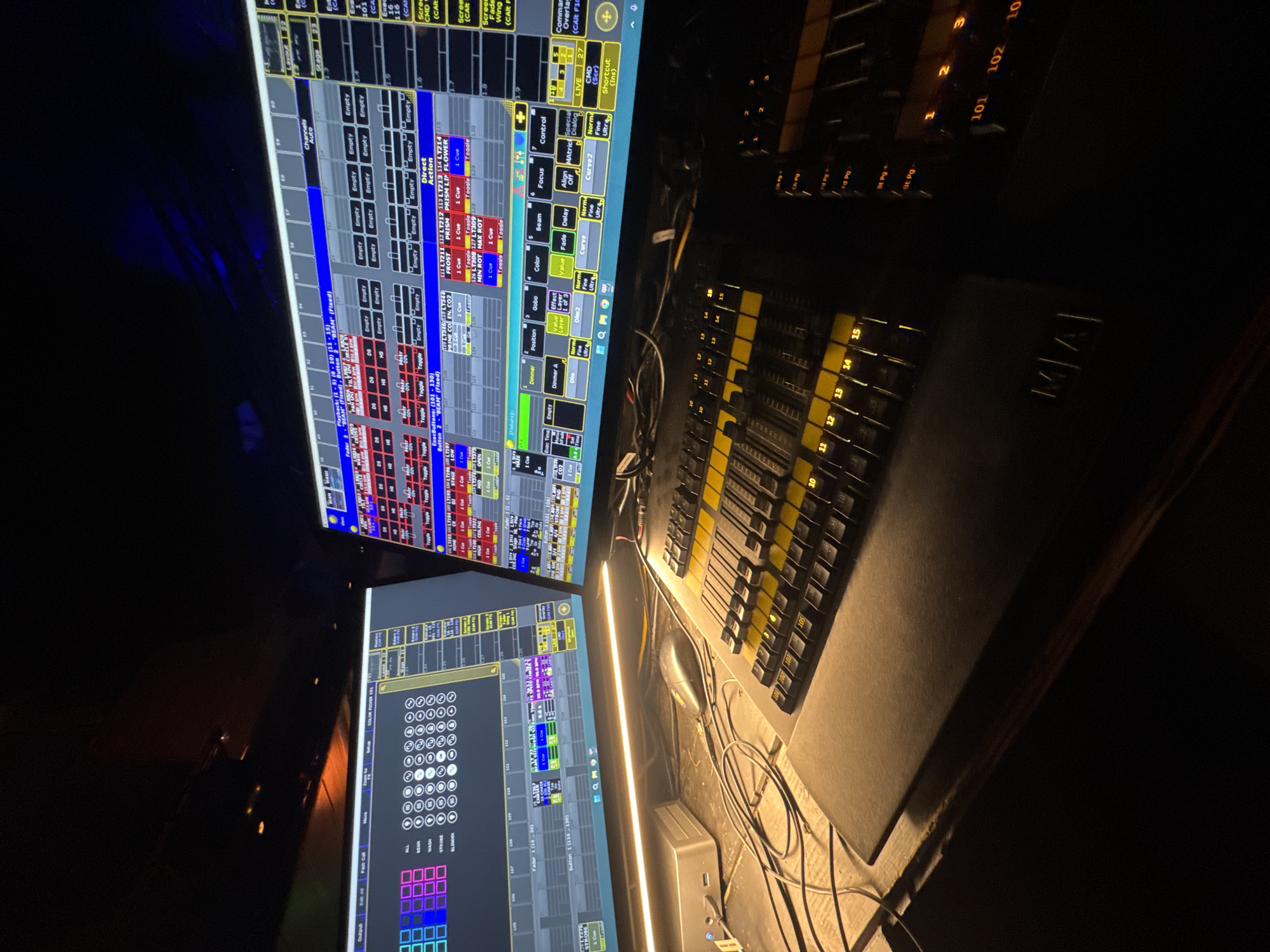 The image shows a dimly lit room, likely a control booth for a theater or event space, featuring two large computer monitors displaying complex lighting control software. The monitors are populated with grids of squares, sliders, buttons, and text, indicating a high level of technical operation. In the foreground, a black control console with numerous faders and illuminated buttons is visible. The console has branding that appears to read "MA". A black mouse and a small silver device are situated on the desk to the left of the console. The overall impression is one of a professional lighting setup in operation or ready for use. There are no people visible in the image. The location is not identifiable from the visual information provided.