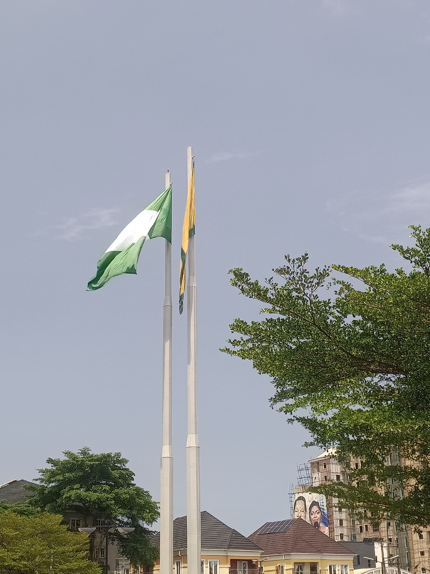 The image shows two flags waving in the wind. The first flag has vertical green and white stripes. The second flag has yellow and green stripes. The flags are on white poles. In the background, there are houses, trees, and a building under construction with a large advertisement. The location is Igboefon, Nigeria, suggested by the presence of the Nigerian flag.