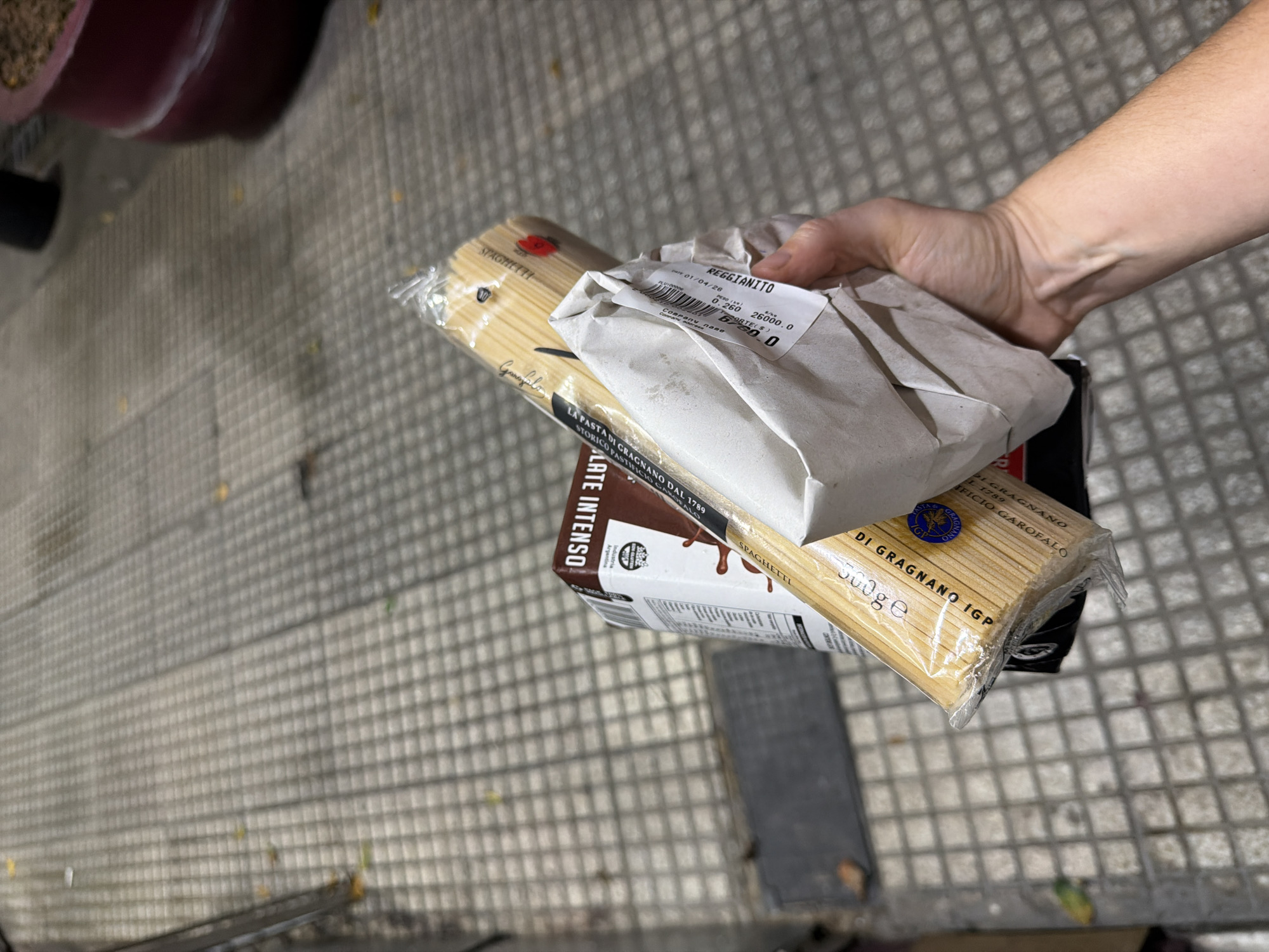 A hand holds a small stack of food items outside on a tiled surface. On top is a package of cheese labeled "REGGIANITO" in a crinkled paper wrapping with a receipt. Below this is a package of Garofalo brand spaghetti, and below that a box of Intenso brand chocolate. The date on the receipt is 01/04/26, and the price is 6760.0. The tiled surface suggests an outdoor area, possibly in Buenos Aires, Argentina, considering the Argentine industry label on the chocolate.