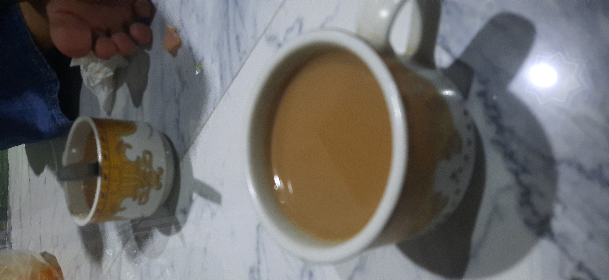The image captures a close-up view of two cups of coffee, one in the foreground and another slightly blurred in the background. The foreground cup is a light brown color with a white rim and a decorative pattern around its base. It is filled with a creamy, light brown liquid, likely coffee with milk. A spoon is partially submerged in the coffee. The background cup is smaller and has a similar decorative pattern. The cups are placed on a marble-patterned surface, possibly a table. In the background, a person's leg wearing blue jeans and a foot with visible toes can be seen. There is also a crumpled white tissue and a small piece of food on the surface. The lighting suggests an indoor setting, possibly a home or cafe, with a bright light source creating reflections on the coffee and surface. The location is specified as Cisauk, Indonesia.