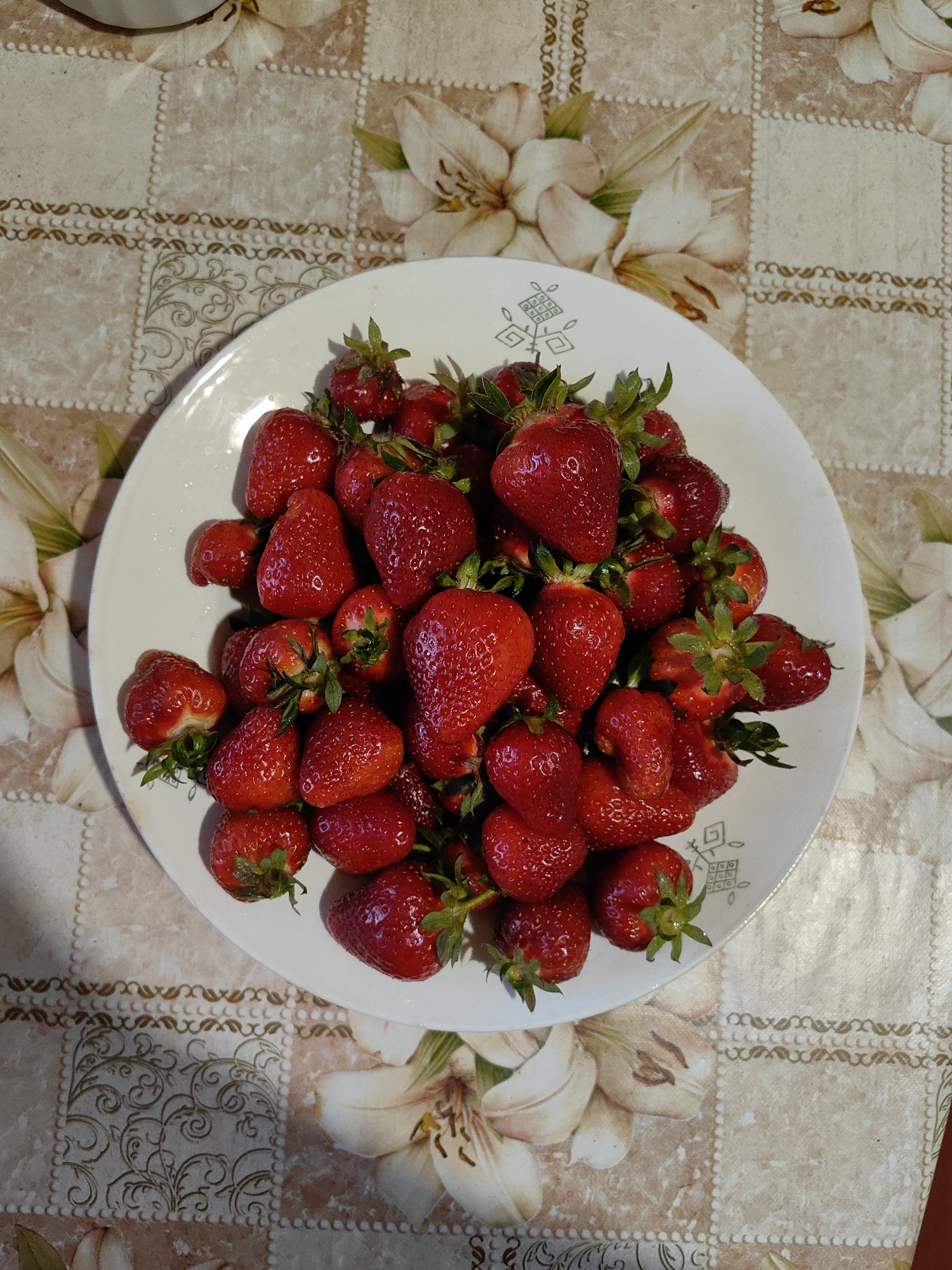 A top-down shot shows a white plate filled with fresh, red strawberries. The strawberries are of varying sizes, with green leafy stems attached. The plate rests on a tablecloth with a repeating pattern of beige squares and stylized lilies. The setting appears to be indoors, likely a kitchen or dining area. The location, Зуйское сельское поселение, Ukraine, is not visually discernable from the image. No people are visible.