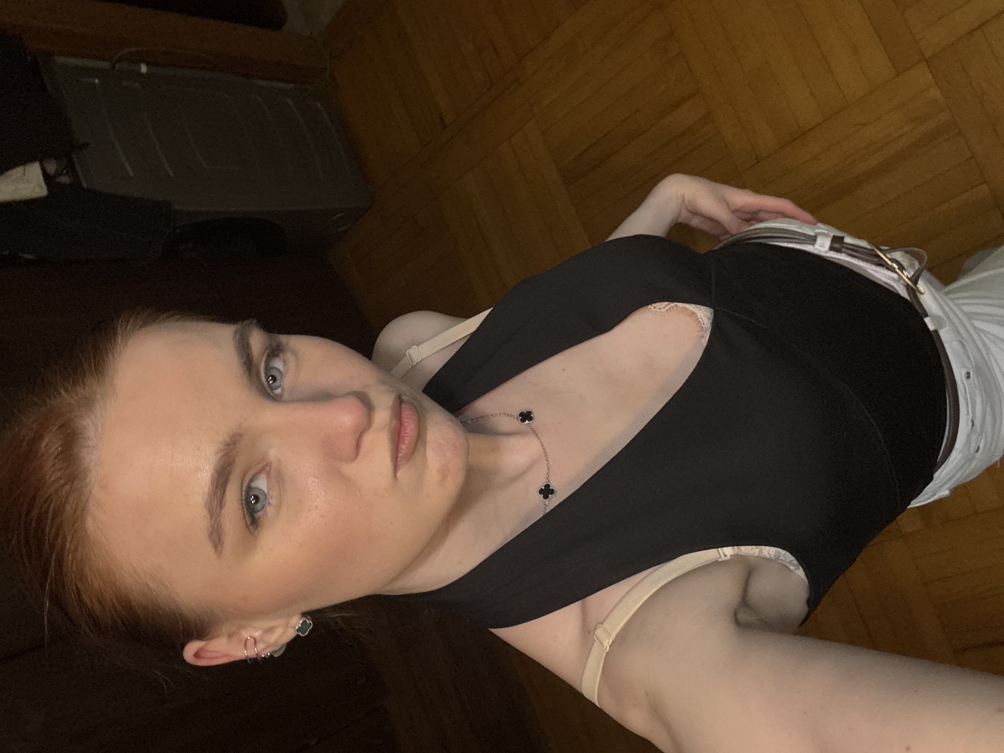 The image is a selfie taken from a low angle, focusing on a young woman with red hair and light blue eyes. She is wearing a black top with a deep V-neck and thin straps, a beige bra is visible under the straps. She also wears a necklace with black clover-shaped pendants. Her outfit also includes a white pair of pants with a brown belt. The background includes a wooden floor and part of a dark-colored furniture. There is no information in the image to suggest the location is Sinyavino, Russia.