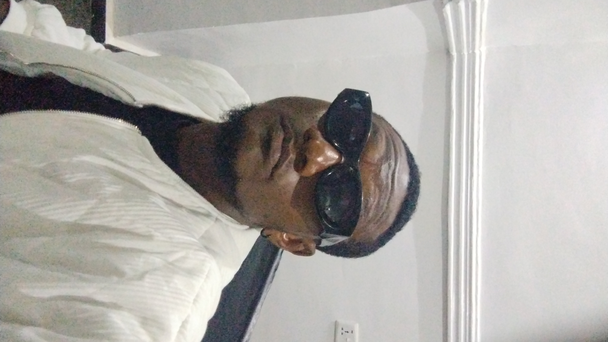 The image shows a Black person with dark sunglasses lying down, possibly in a bed or on a sofa. The person is wearing a white, puffy jacket. The background includes a white wall with a decorative column and an electrical outlet. The location is unknown.